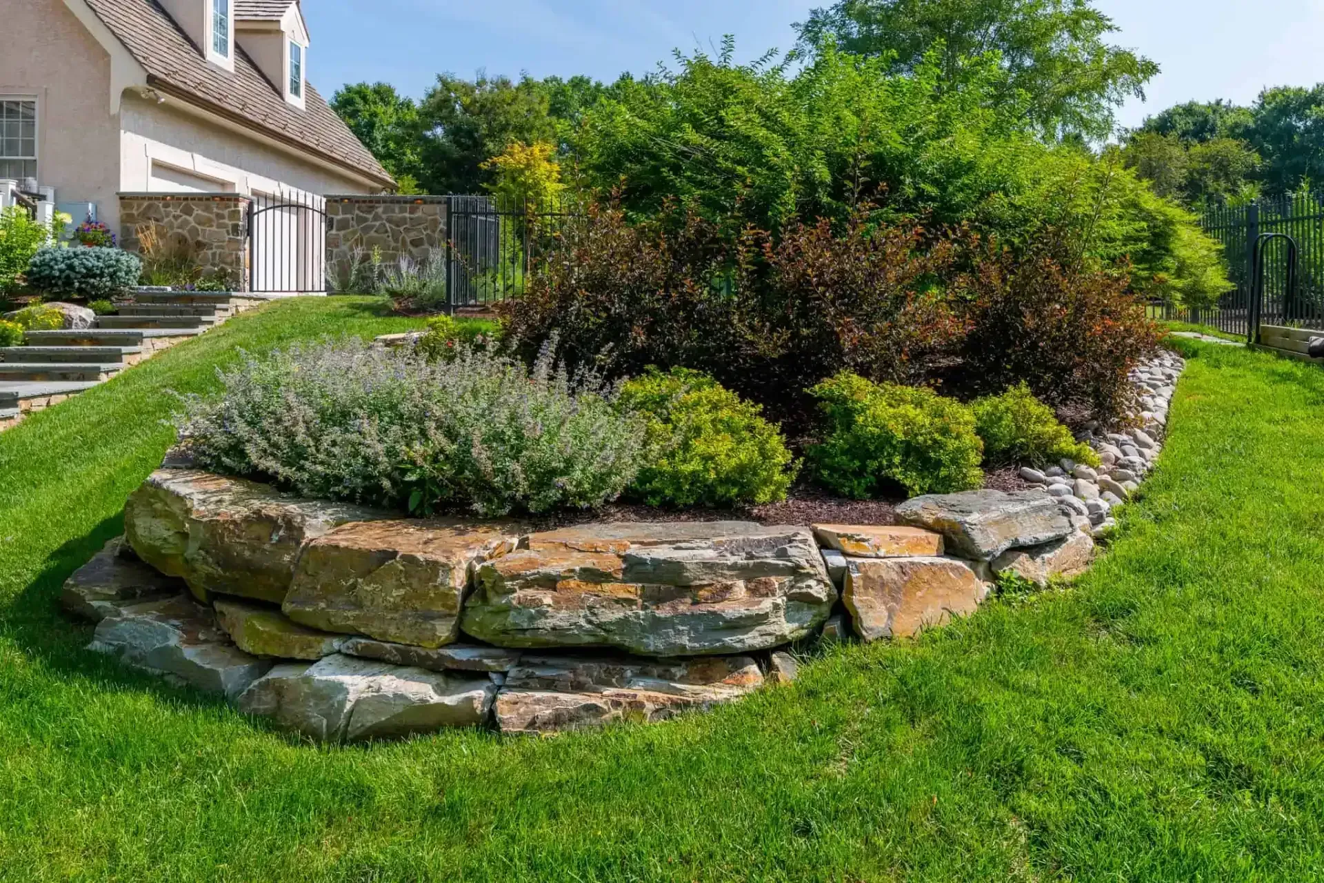 Stone garden bed with layered rocks, shrubs, and green lawn beside a house