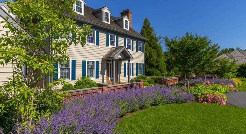 Large colonial house with blue shutters, surrounded by purple flowers and a manicured green lawn