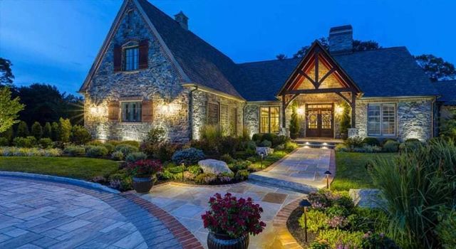 Stone house at dusk with warm lights, landscaped garden, and curved brick driveway.