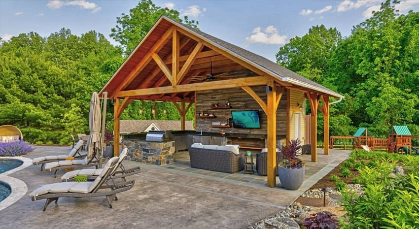 Outdoor stone pavilion with a wood roof, built-in grill, and landscaping in a backyard garden