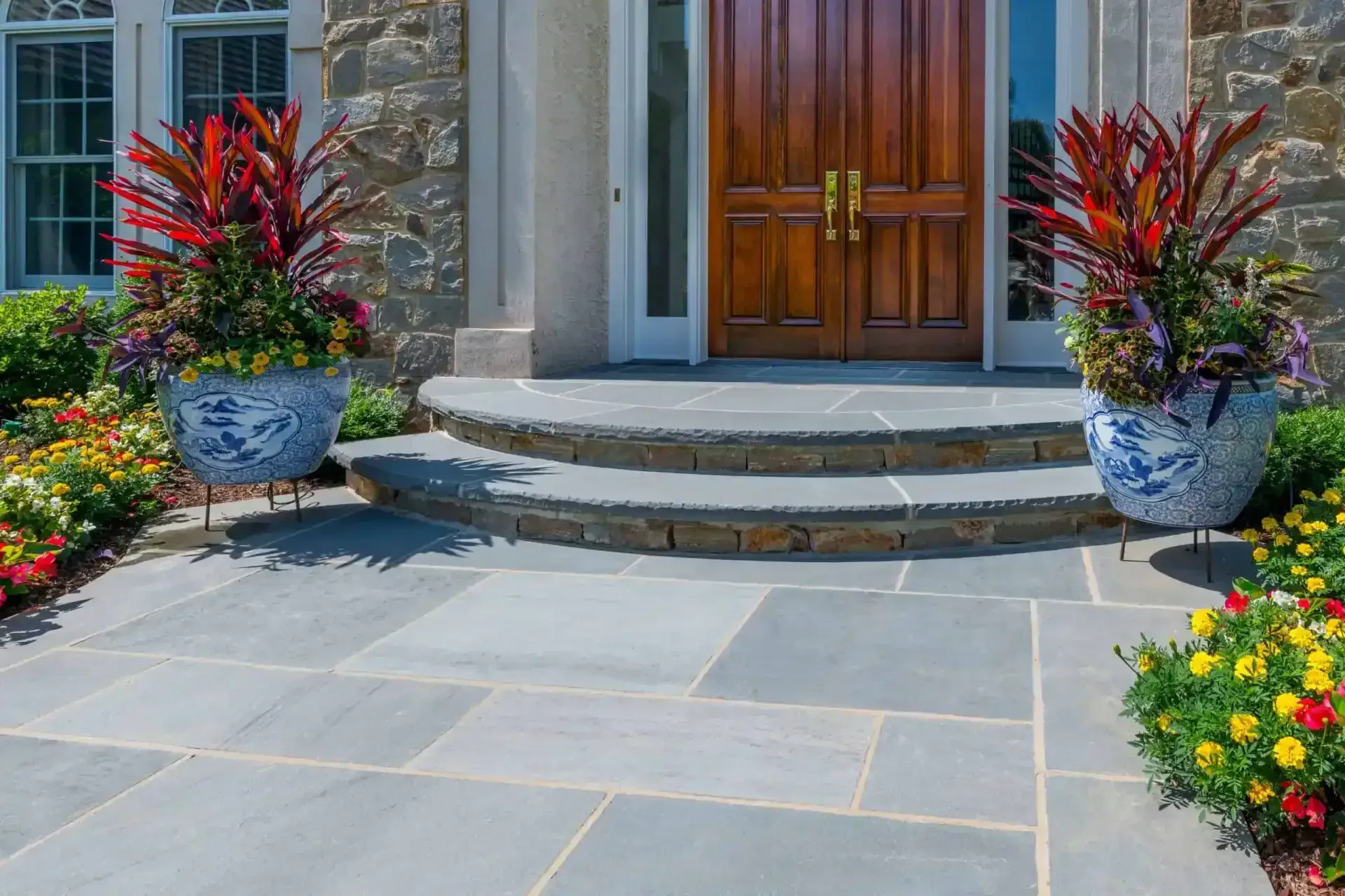 Front entry with wooden double doors, stone steps, and two large potted plants beside a paved walkway