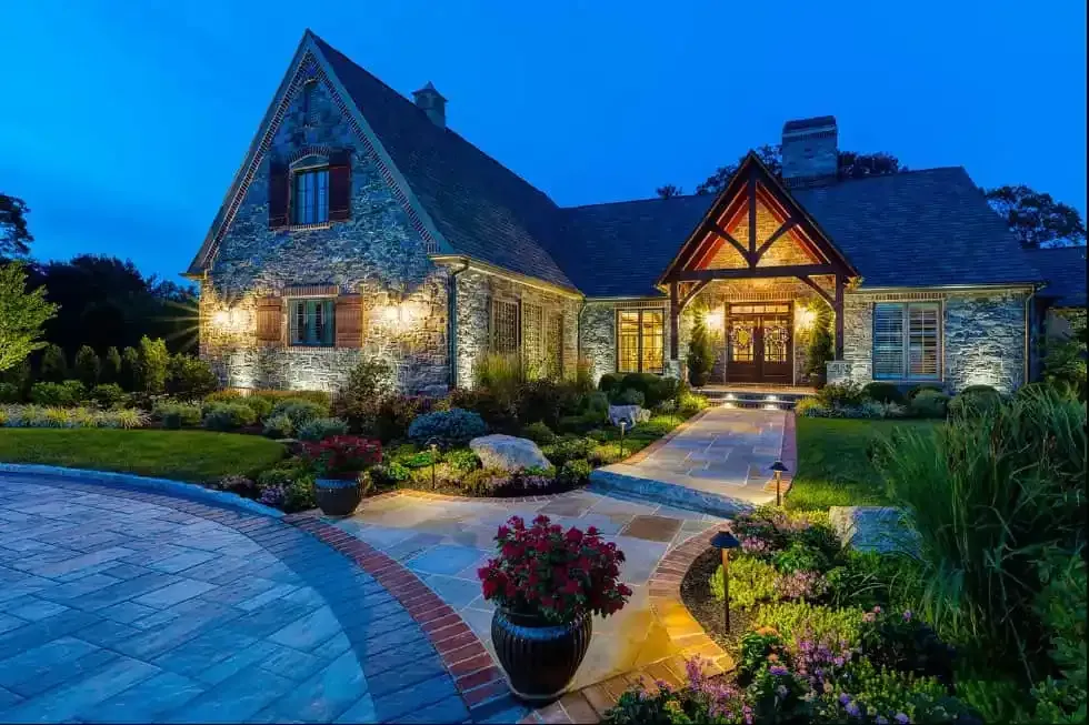 Stone house with lit windows and landscaped garden at dusk, glowing against a blue evening sky