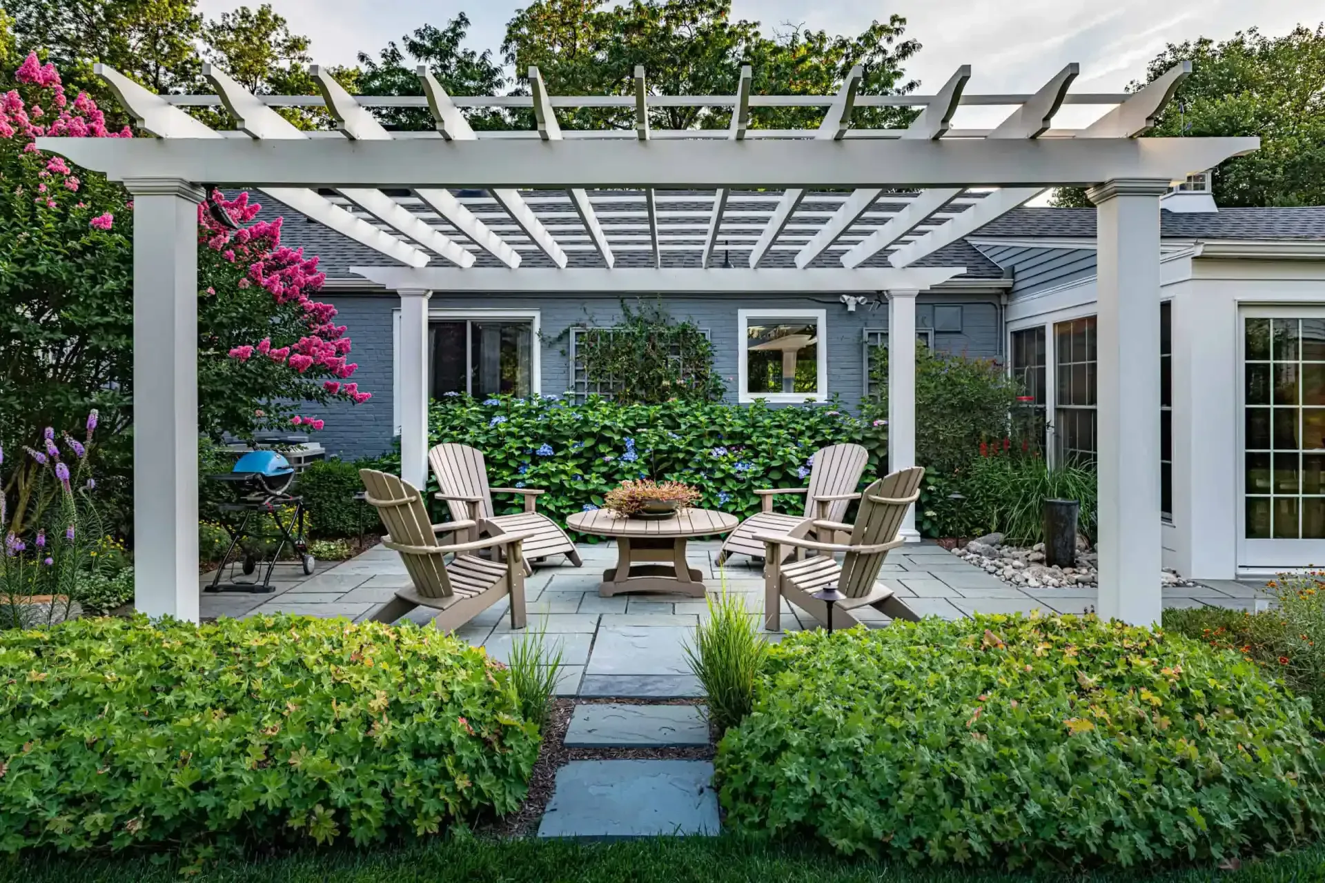 Patio seating under a white pergola beside a house, with lush greenery and pink flowers surrounding it