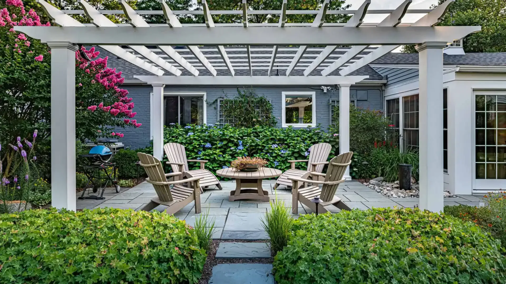 Patio seating area under a white pergola, surrounded by lush greenery and pink flowers.