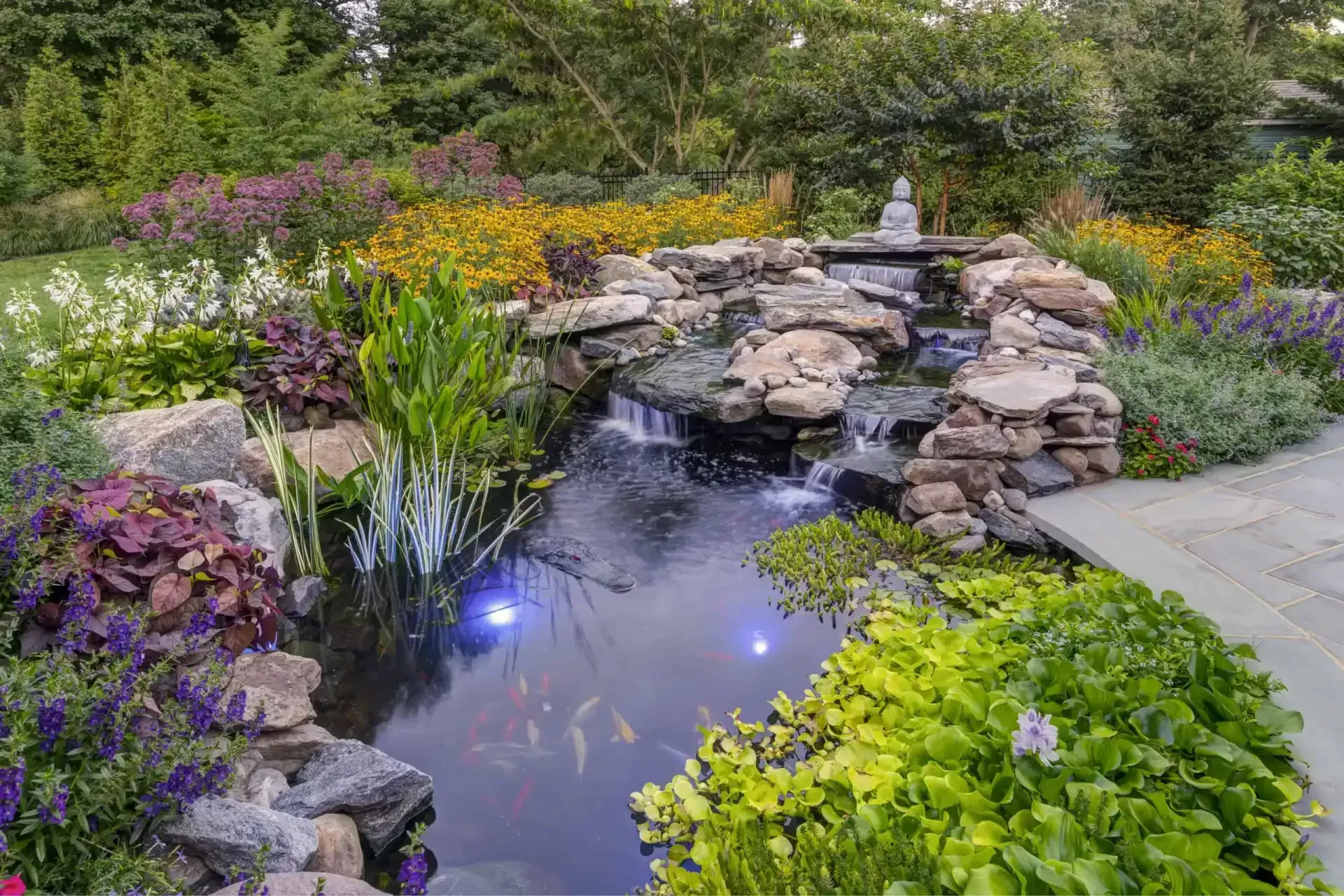 Garden pond with rocks, colorful flowers, and a small waterfall beside a stone path