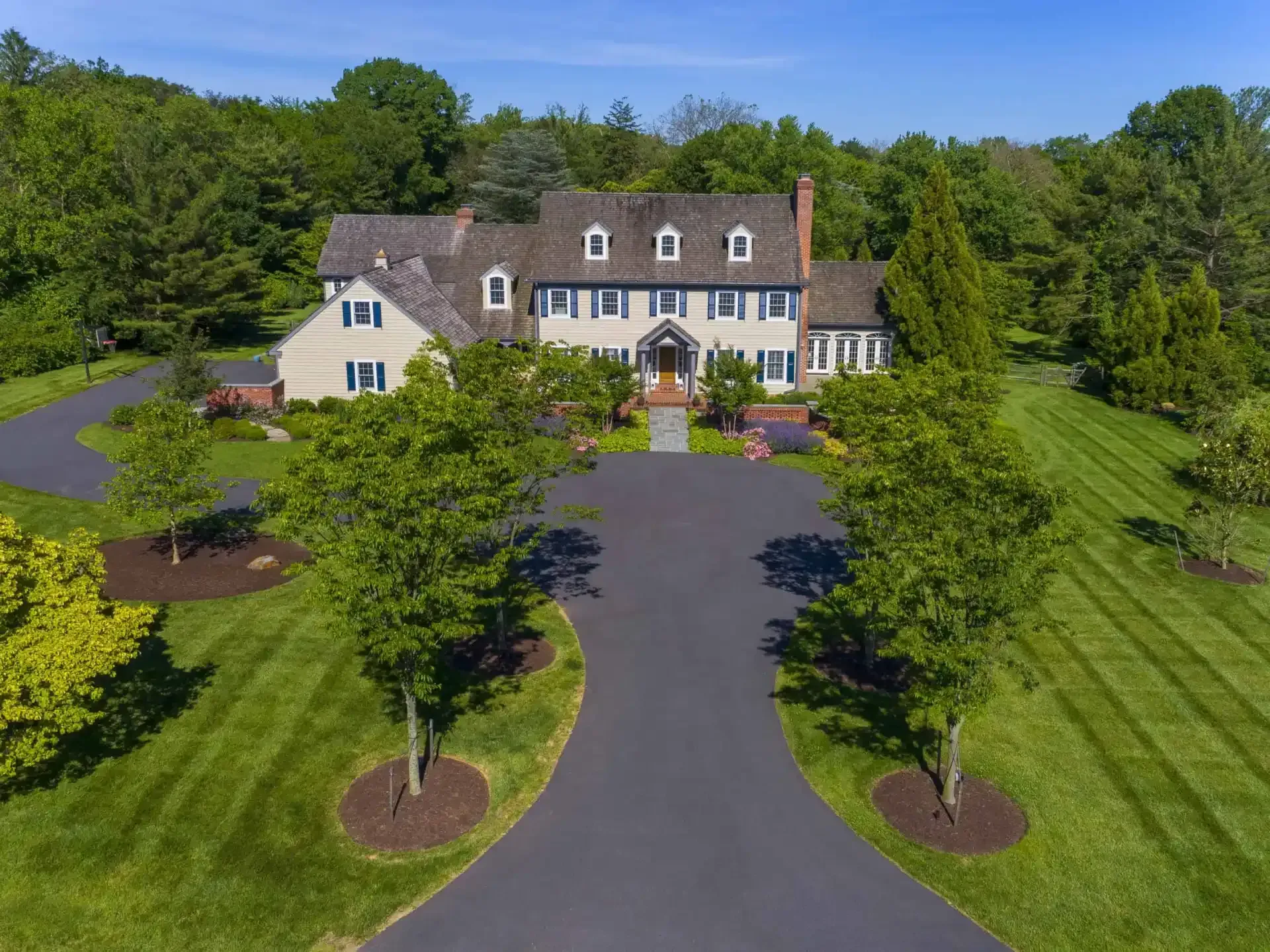 Large suburban house with a dark roof, curved driveway, and manicured lawn surrounded by trees