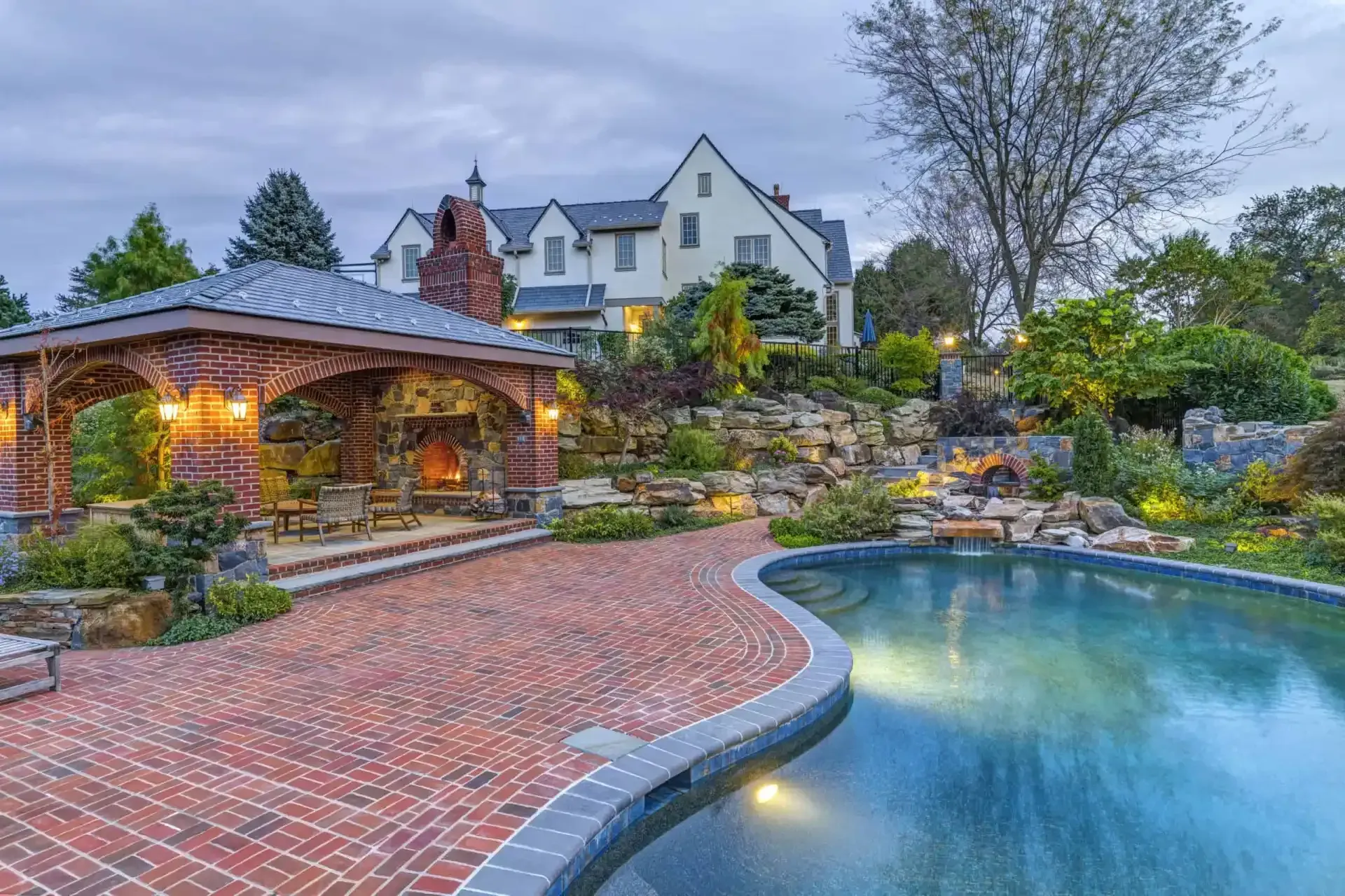 Luxury stone patio with lit gazebo and curved blue pool beside a large house at dusk