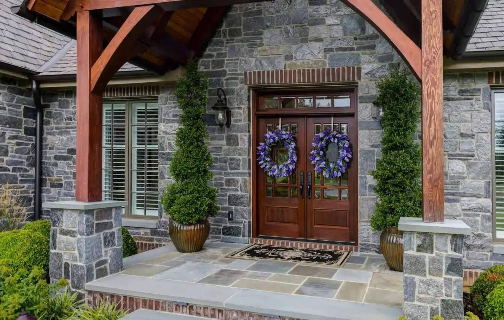 Stone porch with double wooden doors, purple wreaths, and a potted topiary under a timber awning