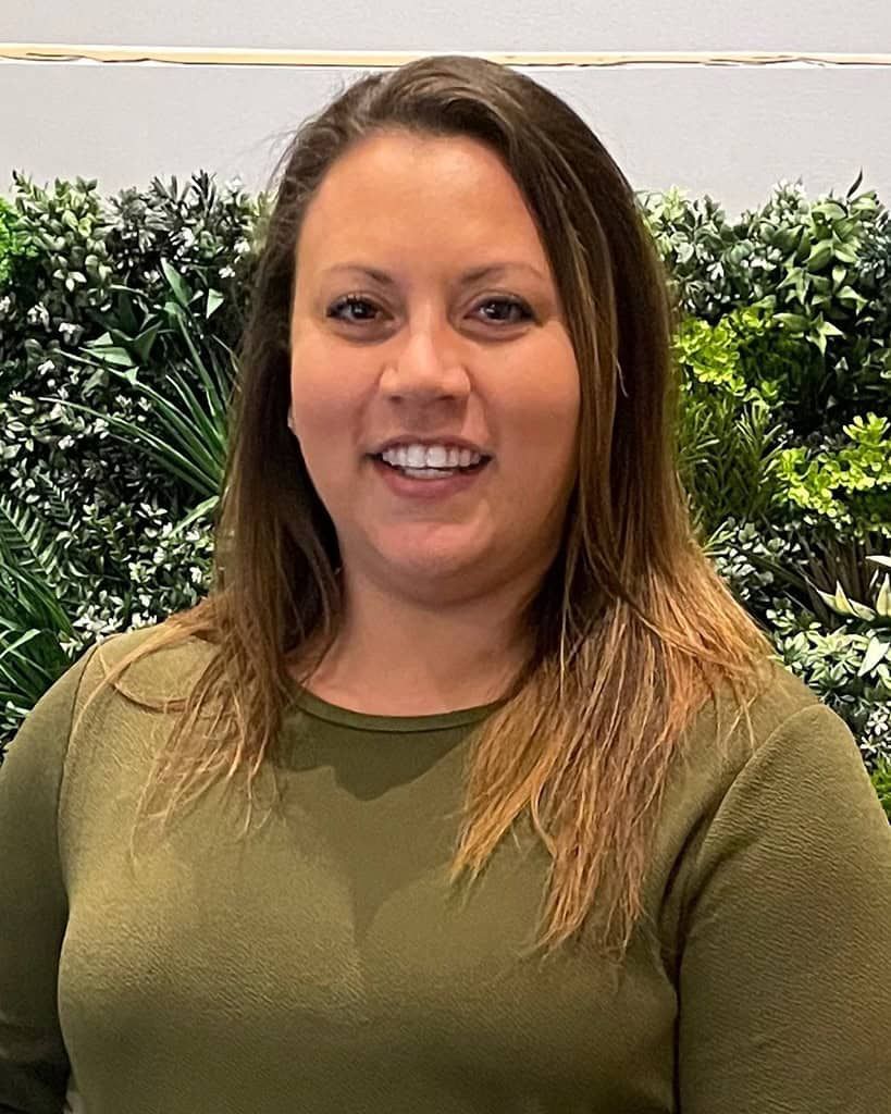 Smiling woman in an olive green top standing in front of a leafy green backdrop