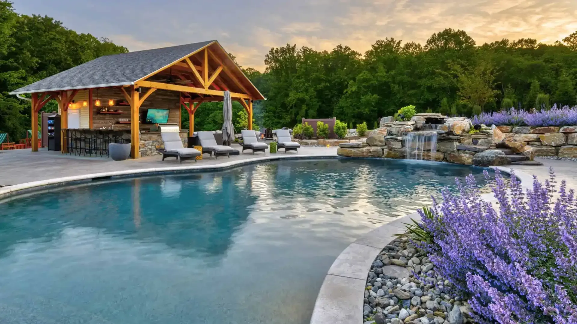 Luxury backyard pool with a wooden pavilion, lounge chairs, stone waterfall, and purple flowers at sunset
