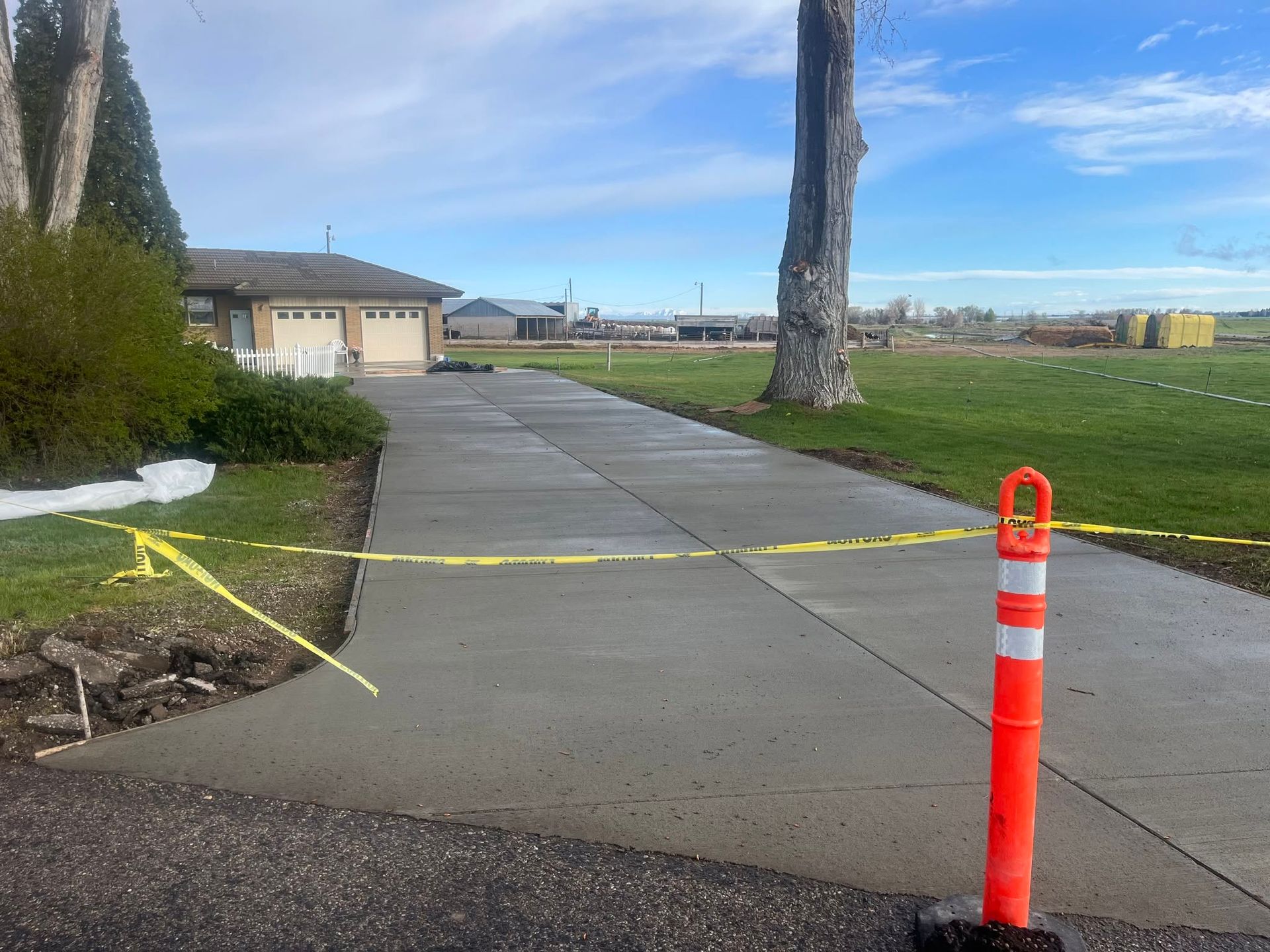 Yellow caution tape blocking a concrete walkway beside a lawn and building, with an orange post in front