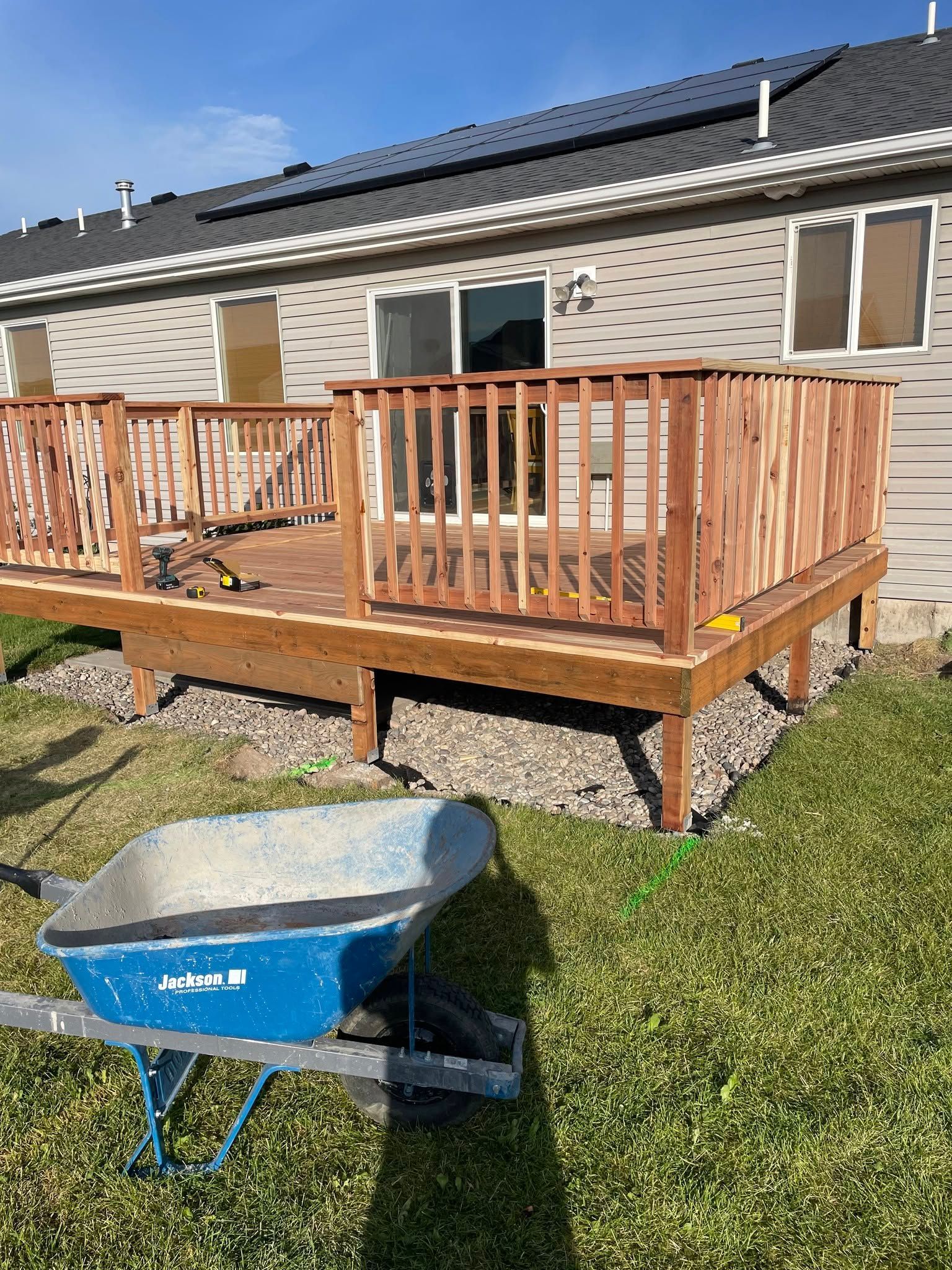 Raised wooden deck with railings beside a house, with a blue wheelbarrow on the lawn.