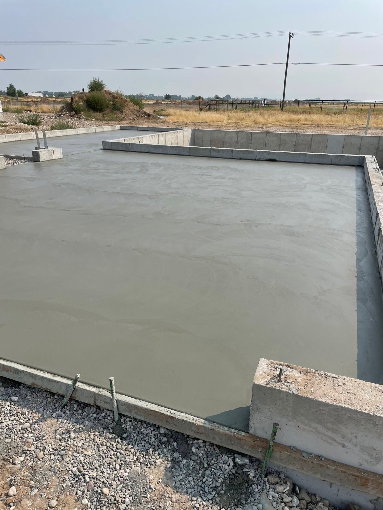 Fresh concrete foundation slab at a construction site with gravel, low walls, and open field beyond.
