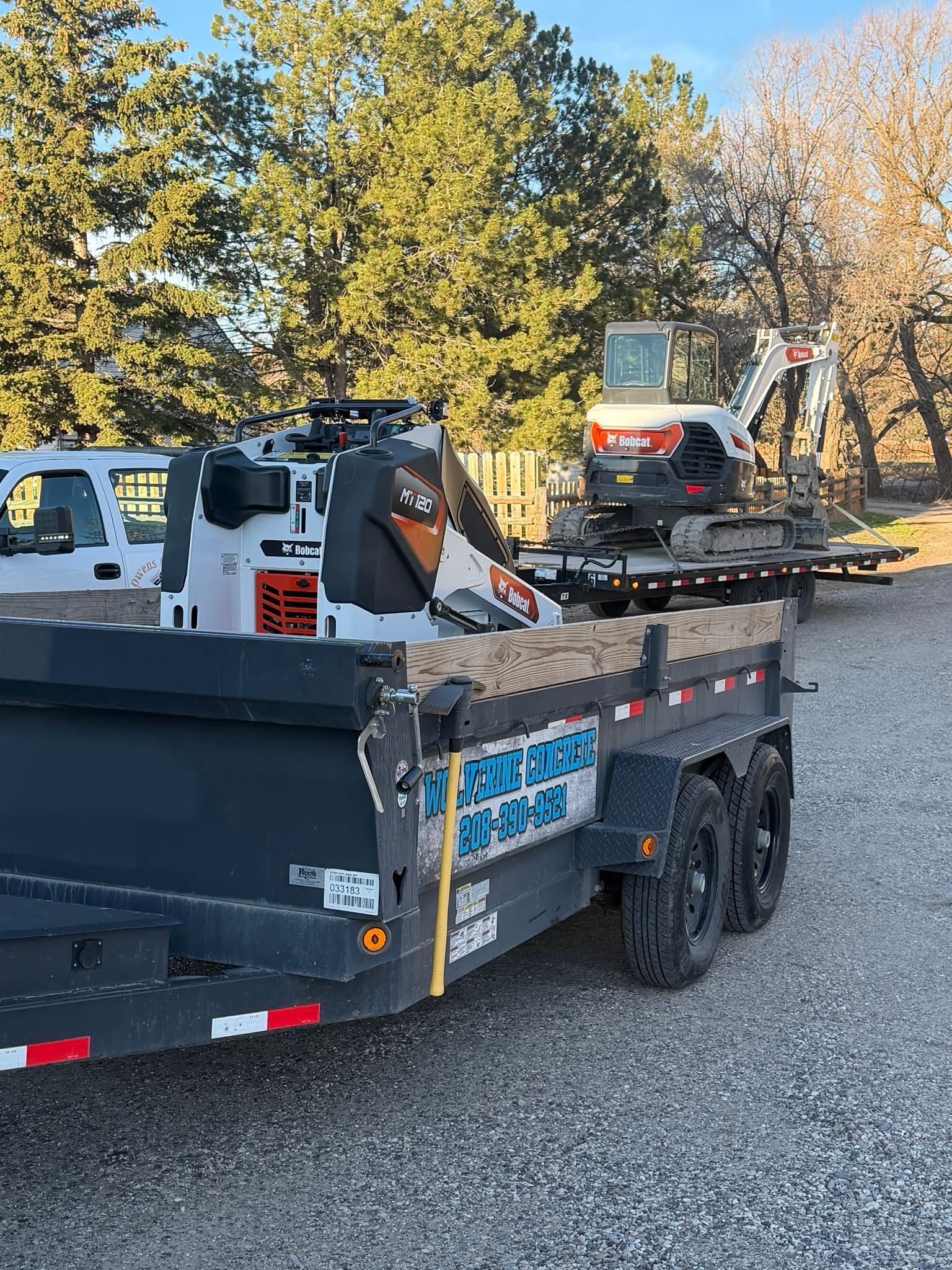 Skid-steer loader on a flatbed tow truck in a gravel lot with trees in the background