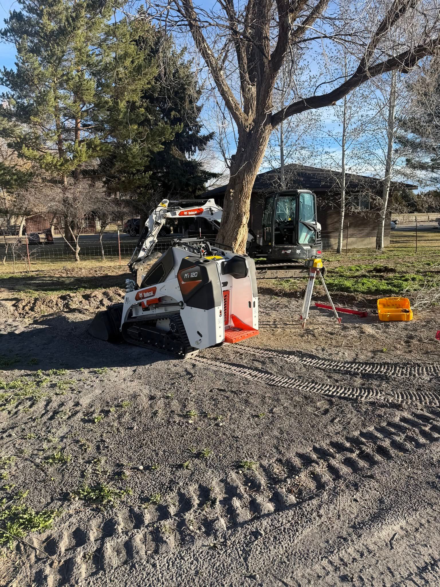 Construction equipment on gravel beside a tree in a sunny outdoor worksite.