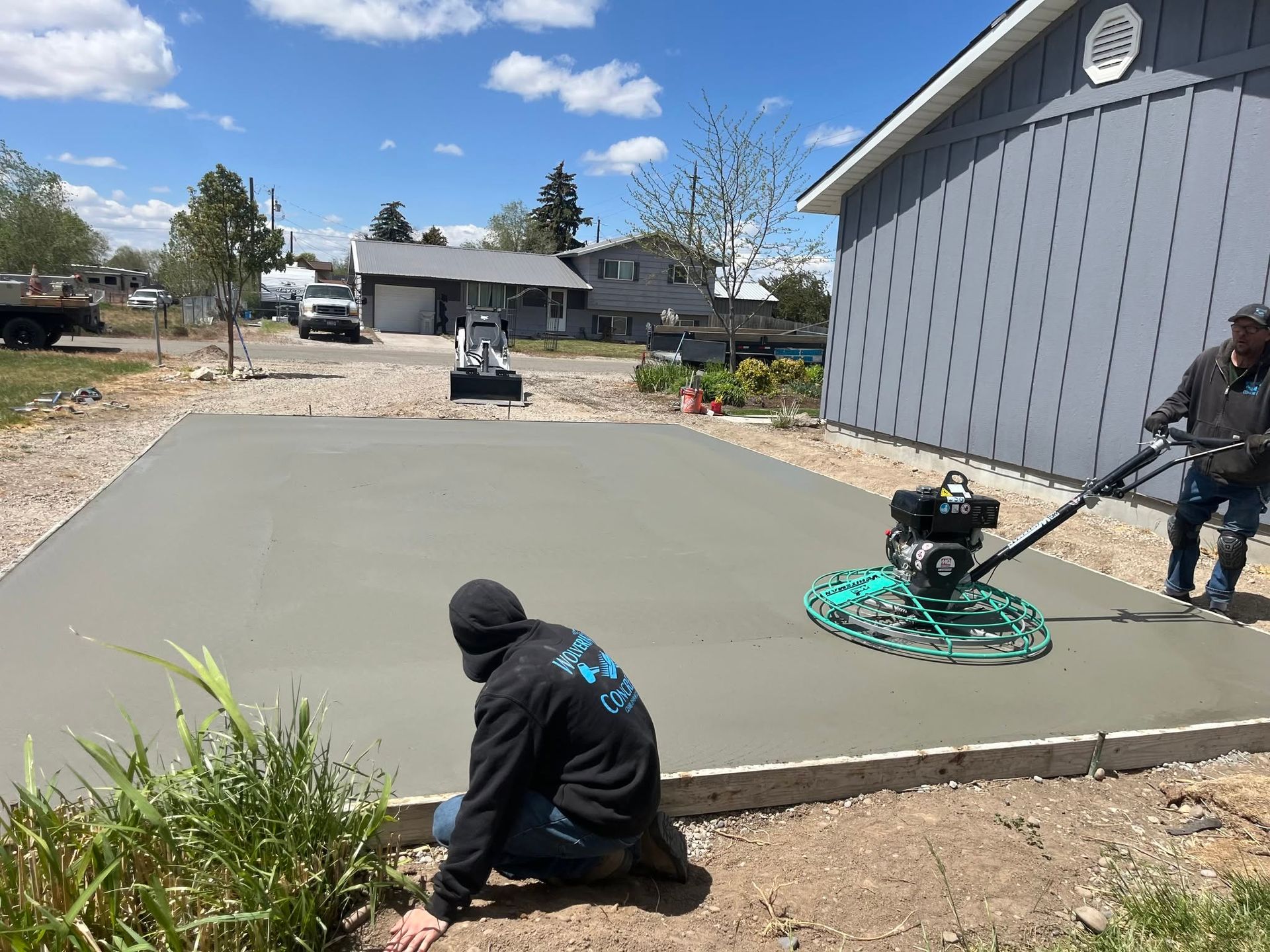 Workers smoothing fresh concrete in a backyard beside a gray house, with tools and hose visible.
