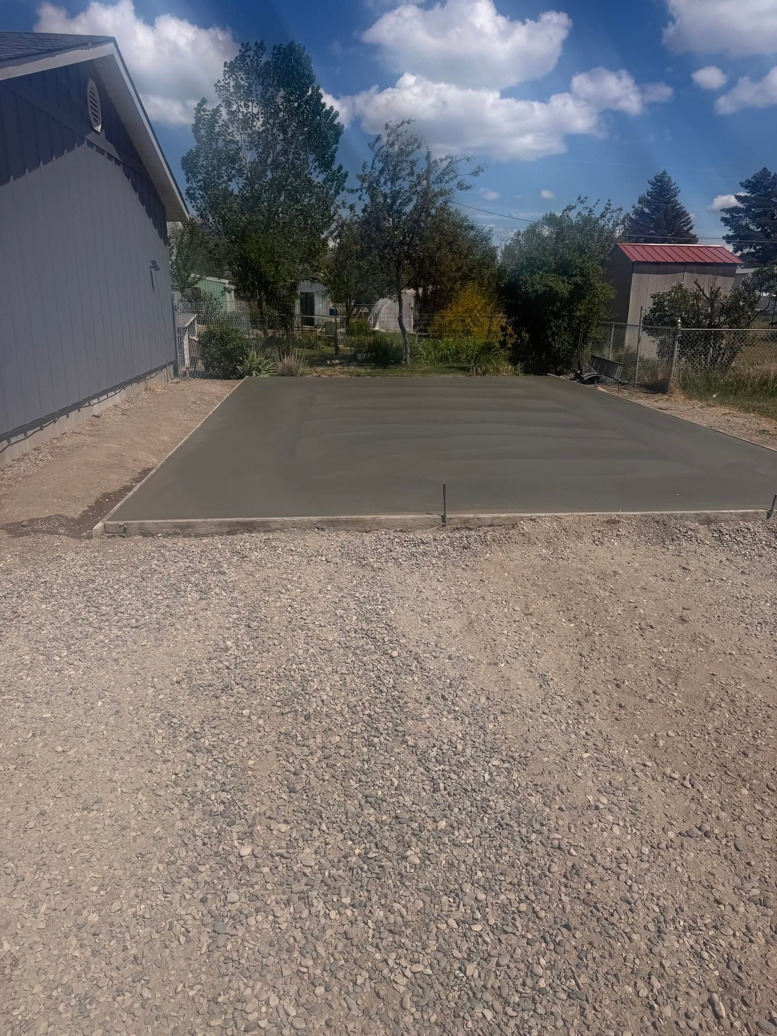 Gravel driveway beside a house, with a concrete pad, trees, and a blue sky with clouds.