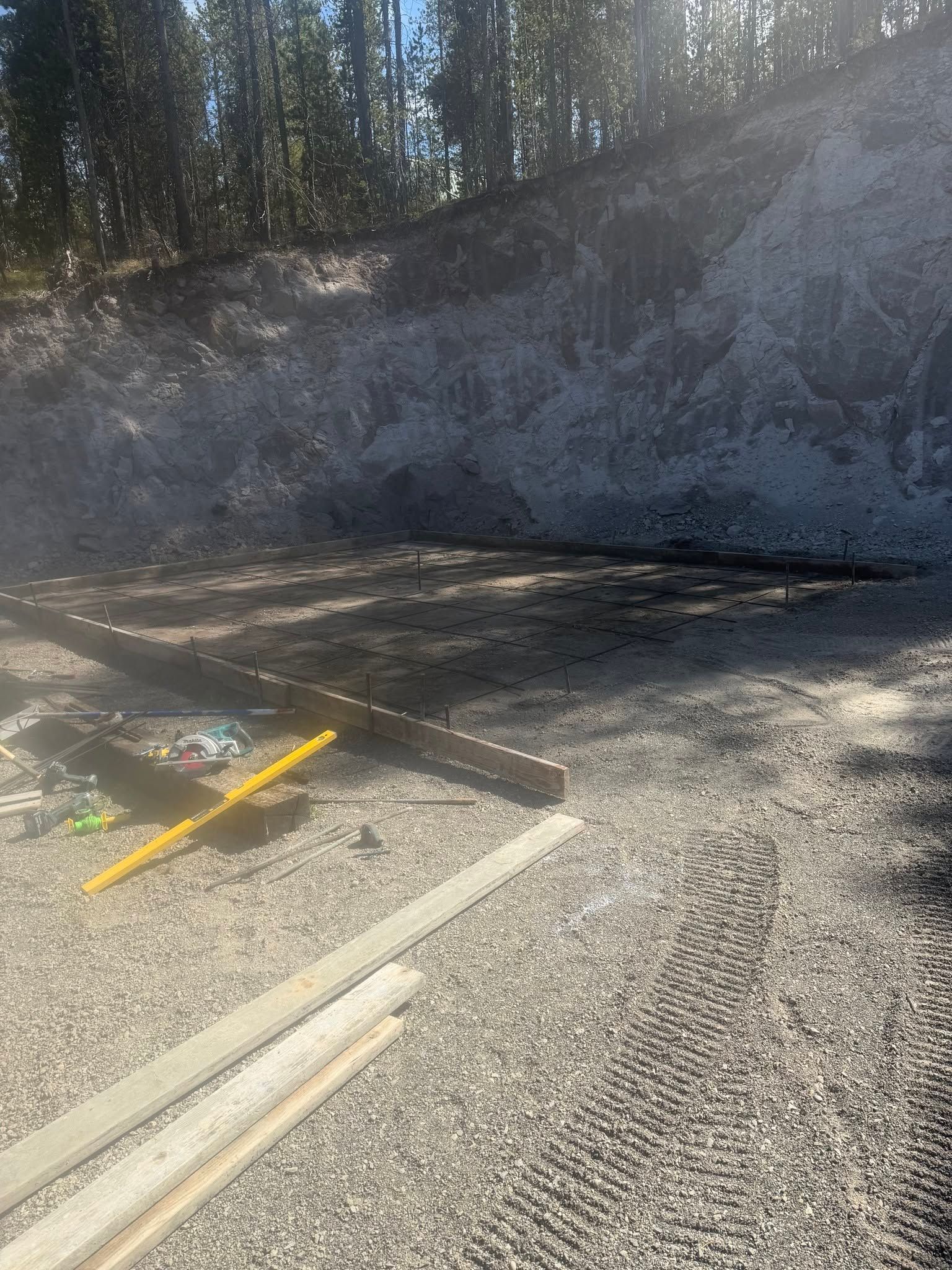 Gravel lot with wooden planks and yellow markings beside a rocky embankment and trees in sunlight
