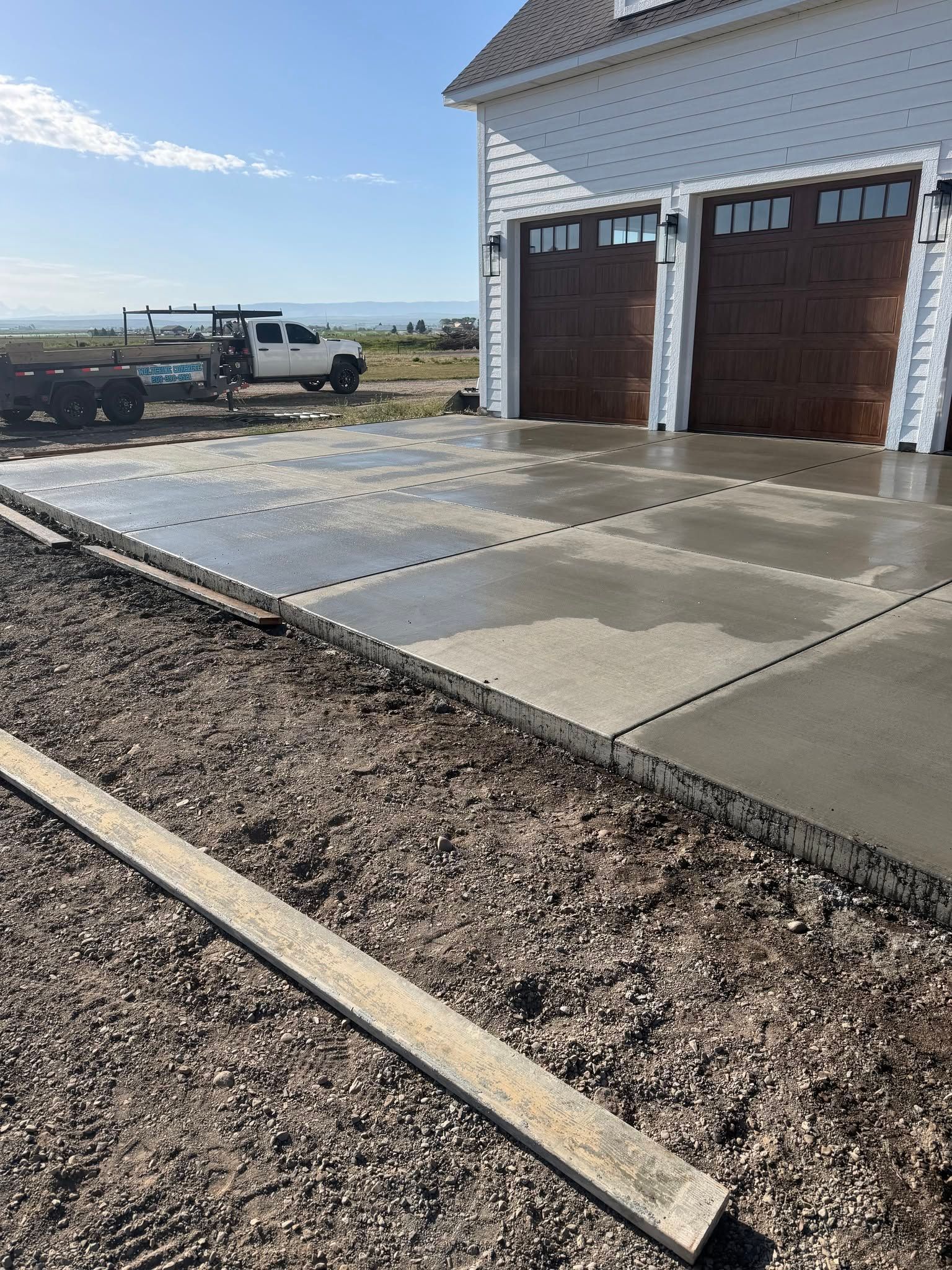 Concrete driveway beside a white house with two brown garage doors, gravel border, and a white pickup truck nearby