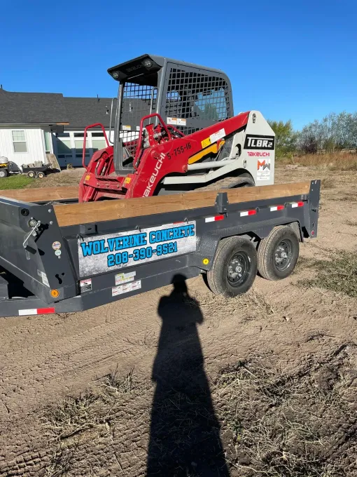 Red skid steer loader on a black utility trailer in a dirt lot under a blue sky