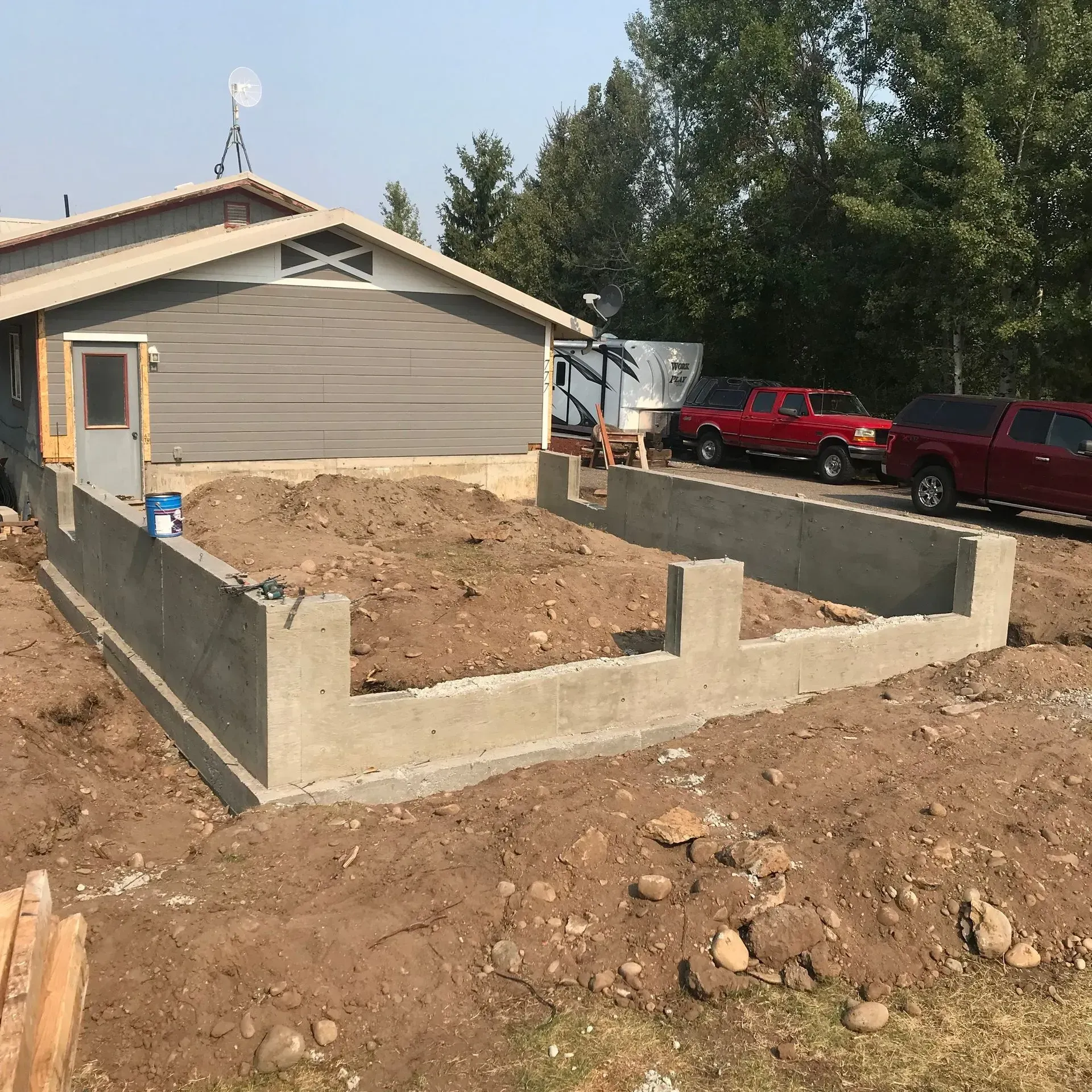 House under construction with new concrete foundation walls and dirt lot in front, trees and parked trucks nearby