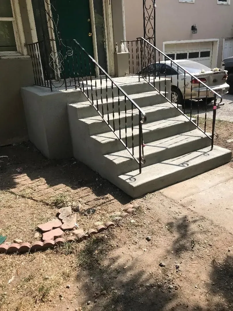 Concrete front steps with black railings leading to a green door, beside a parked car and dry yard.