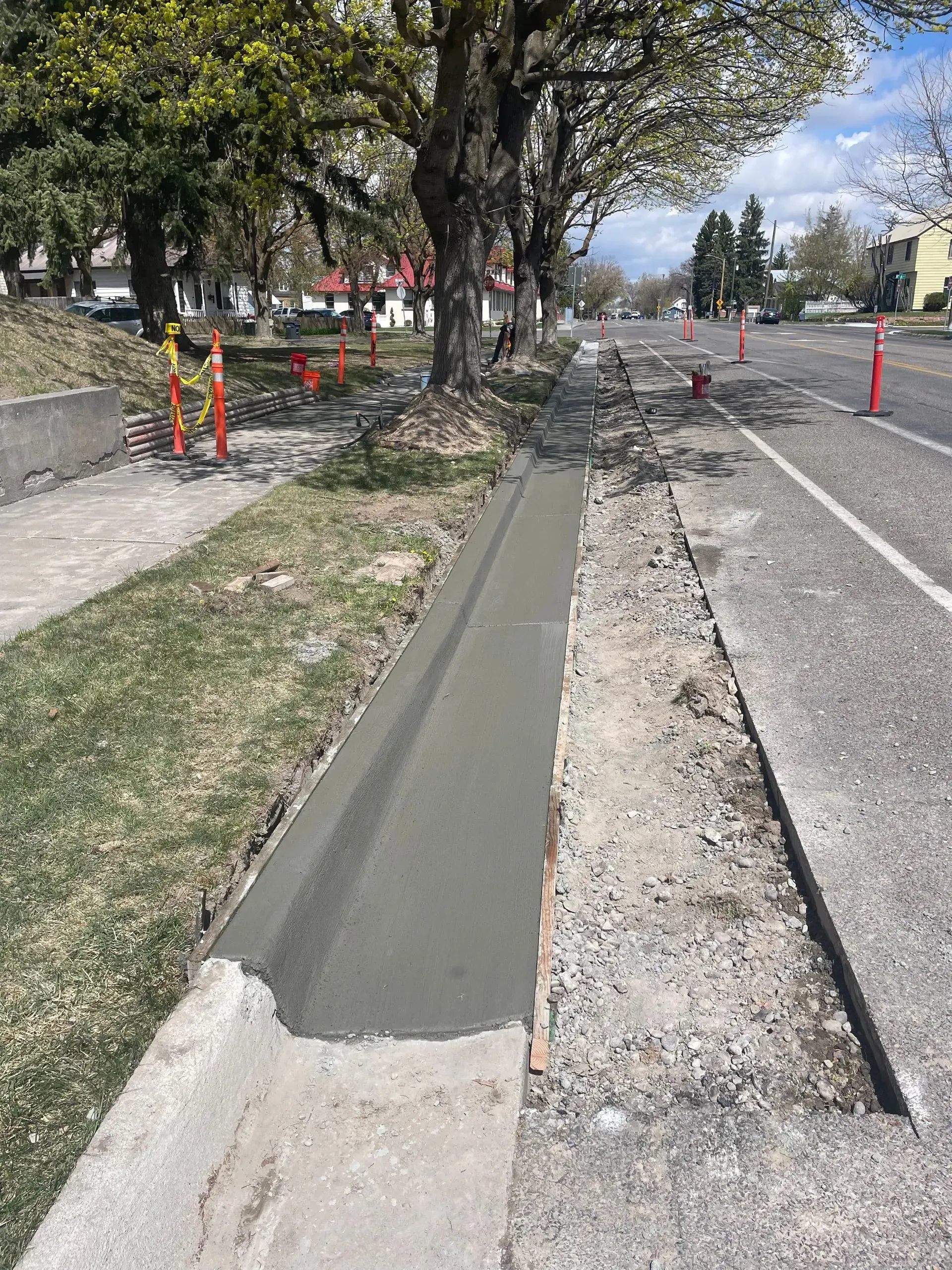 Freshly poured curb ramp beside a street, with orange cones and trees along the sidewalk.