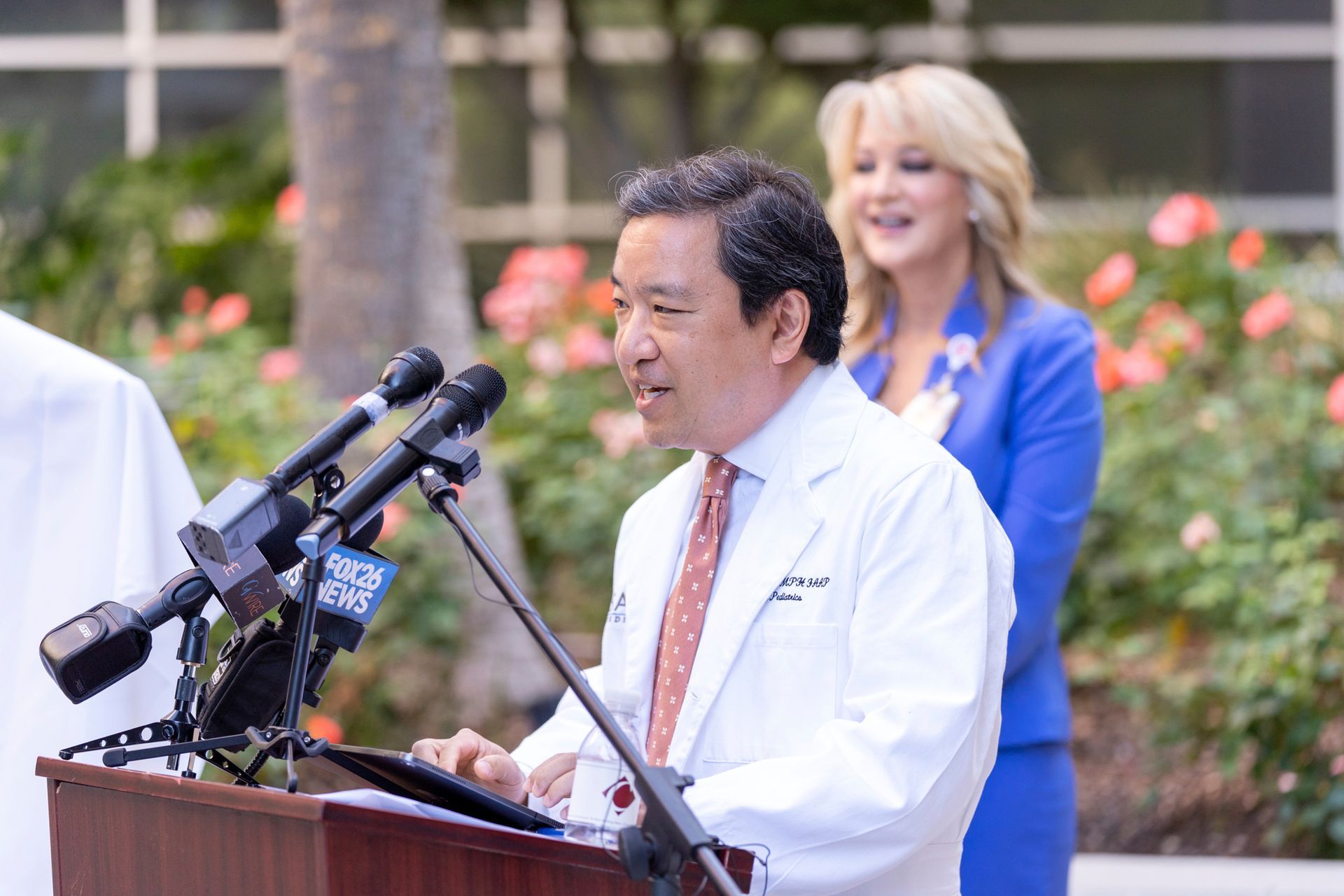 A man in a white coat and tie is standing at a podium with microphones.