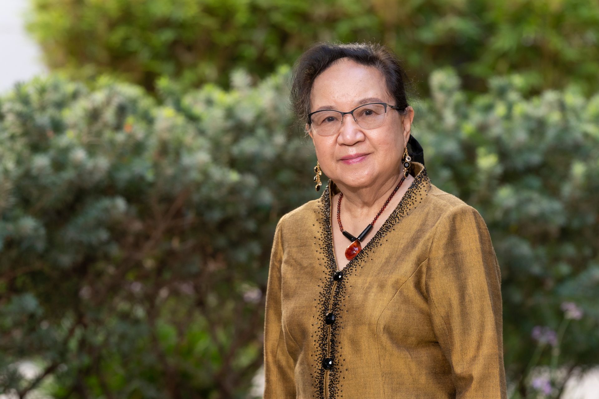 A woman wearing glasses and a necklace is standing in front of a bush.