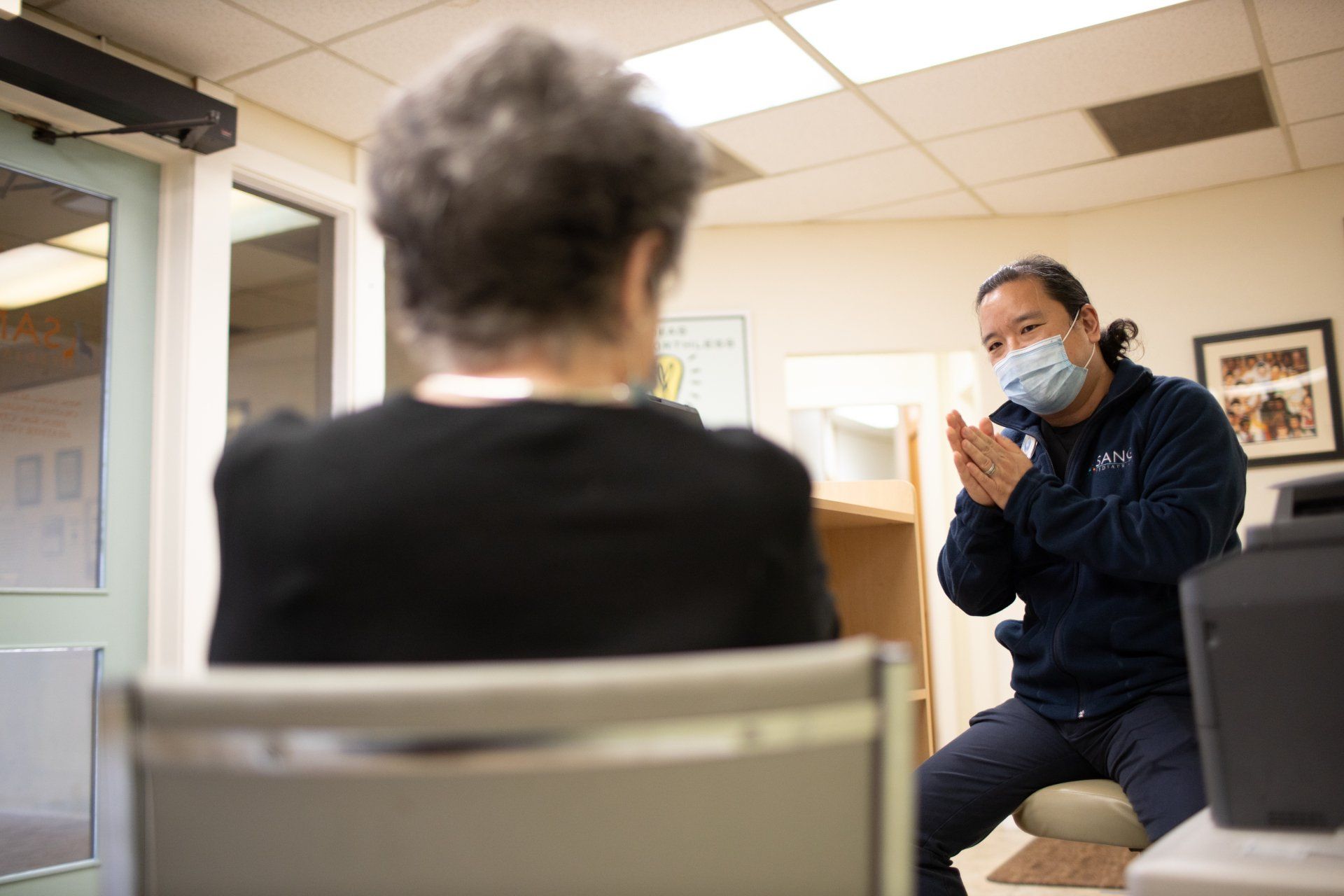 A man wearing a mask is talking to a woman in a chair.