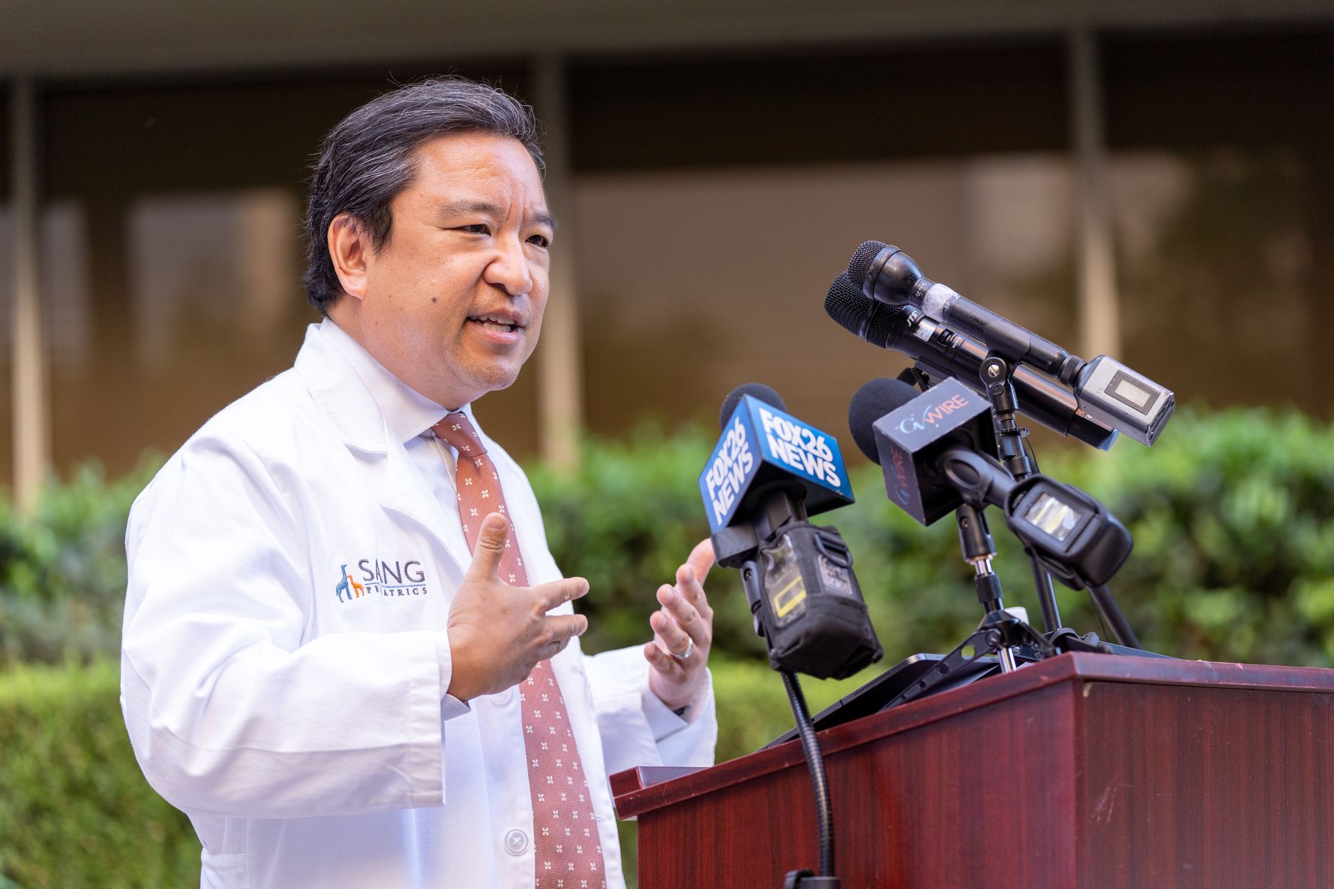 A man in a white coat and tie is standing at a podium talking into microphones.