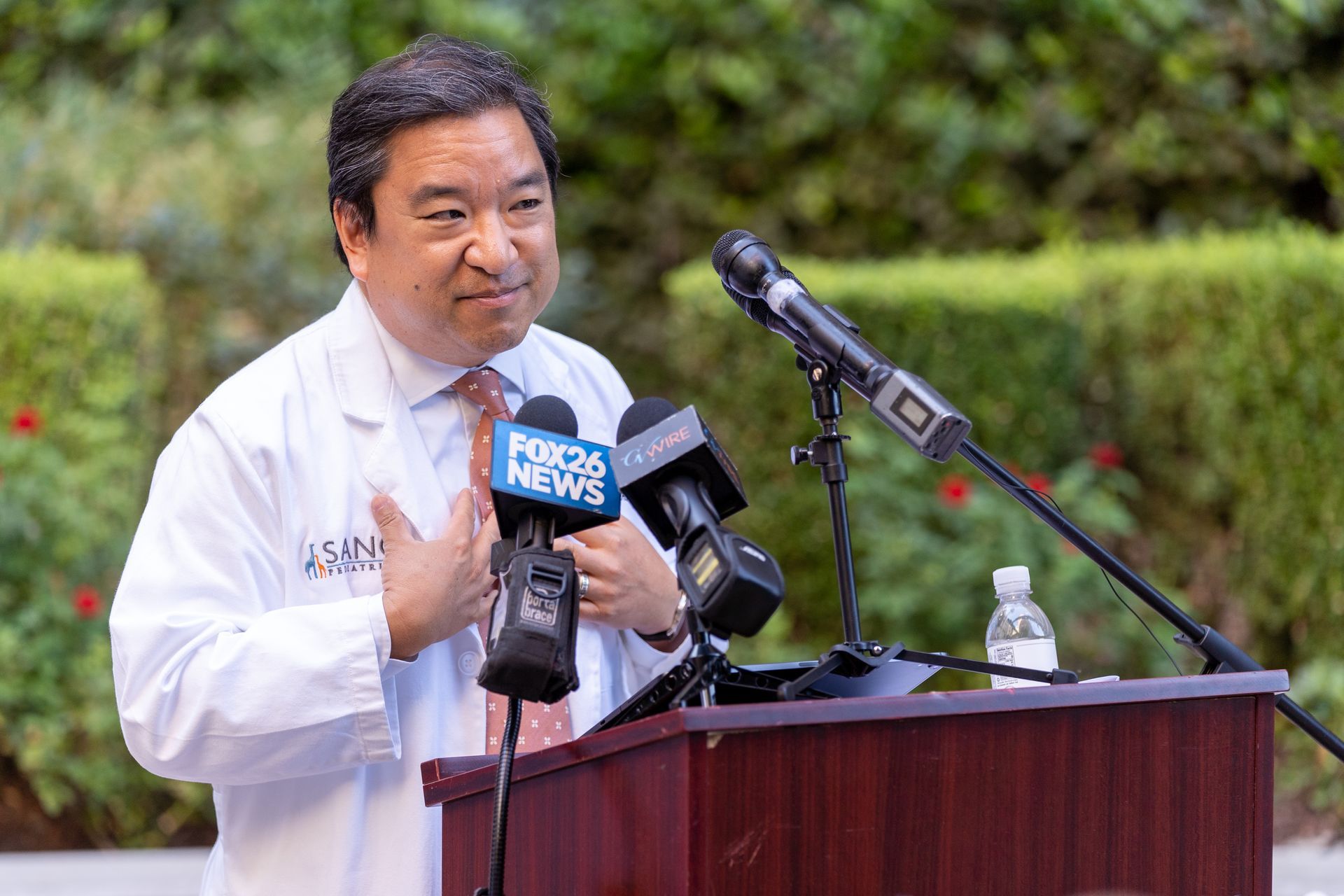 A man in a white coat and tie is standing at a podium holding a microphone.