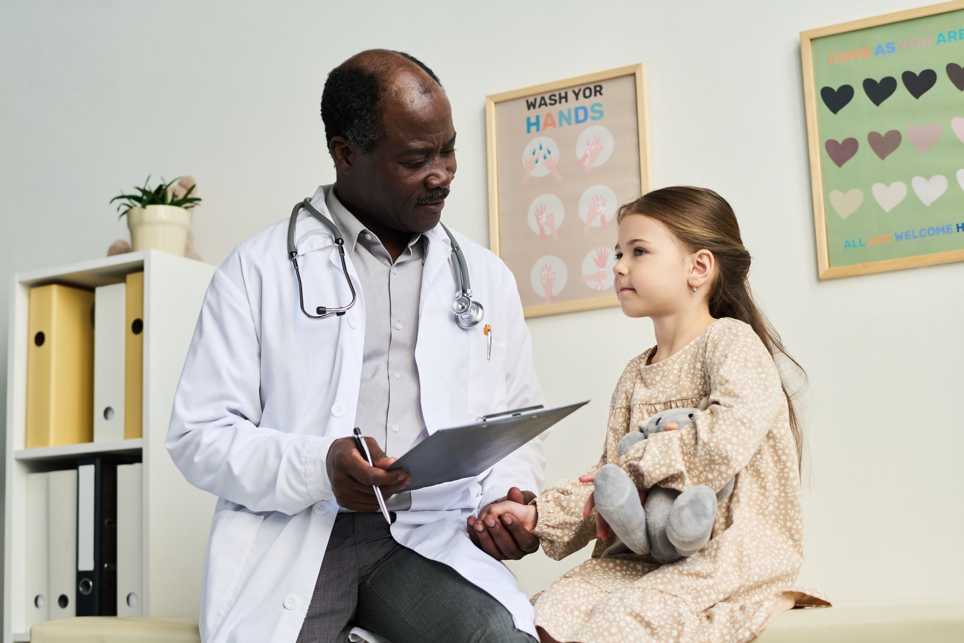 A doctor is talking to a little girl who is holding a teddy bear.