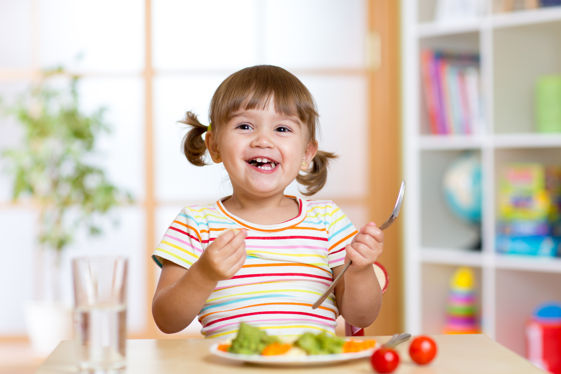 A smiling toddler eating vegetables at a table, learning healthy lifestyle habits during mealtime.