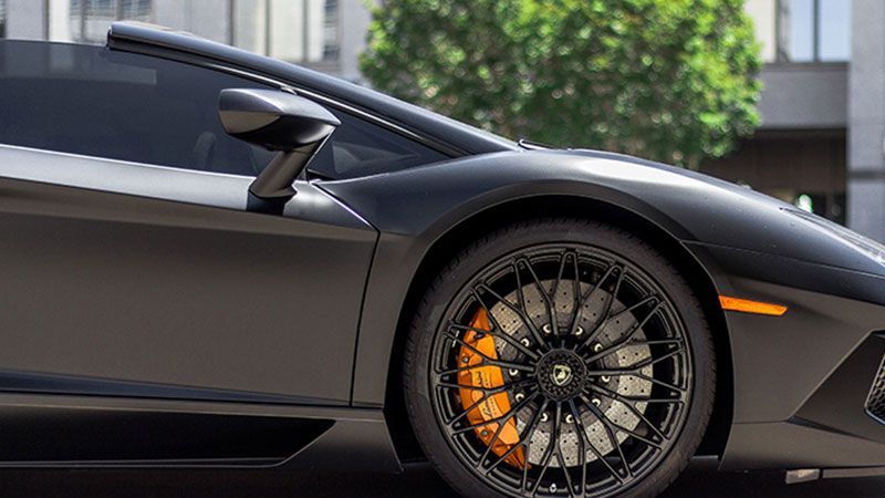 Black Lamborghini sports car, orange brake calipers, black rims, parked outside.
