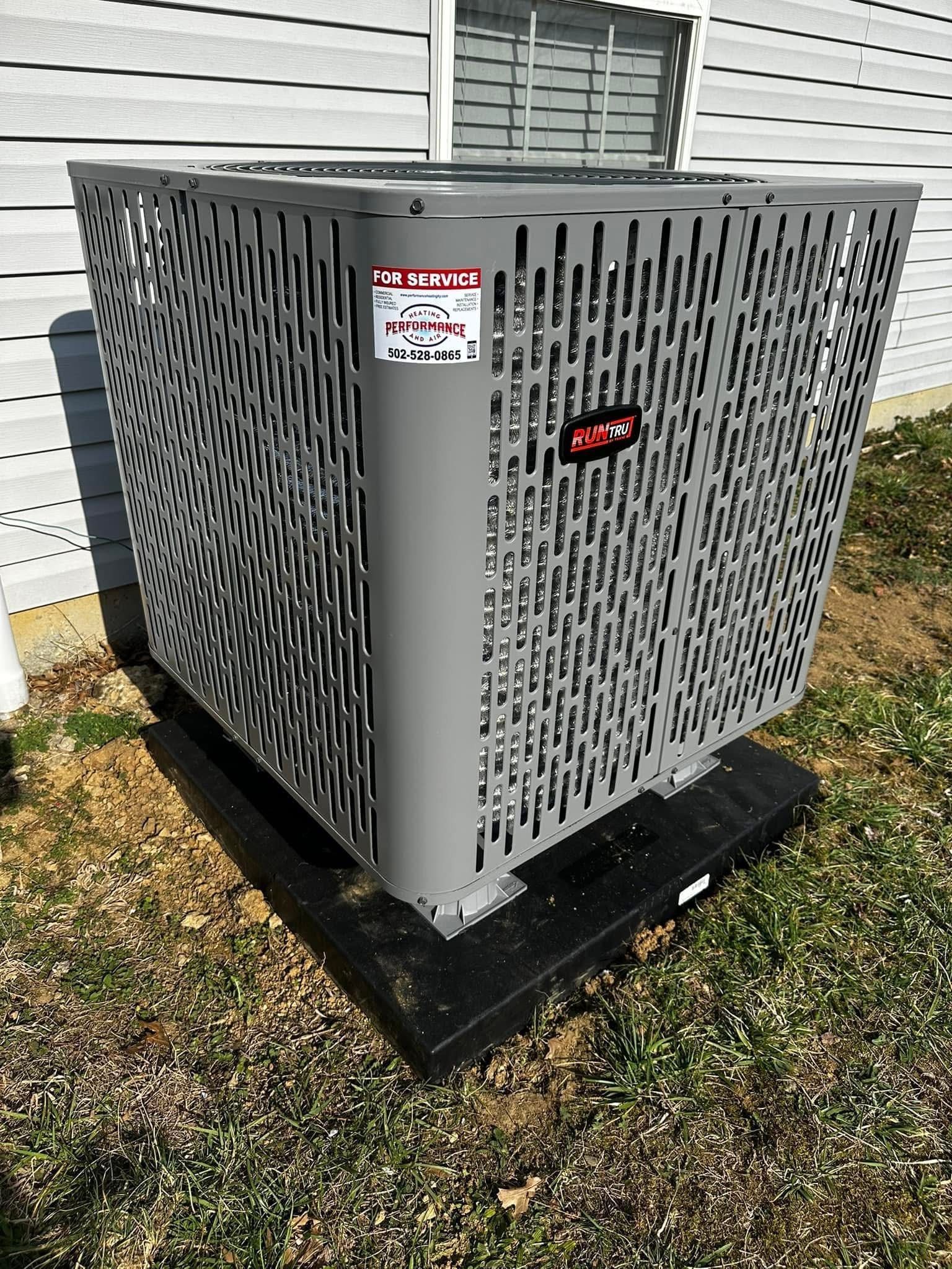 Gray HVAC condenser unit on a black pad beside a house with white siding.