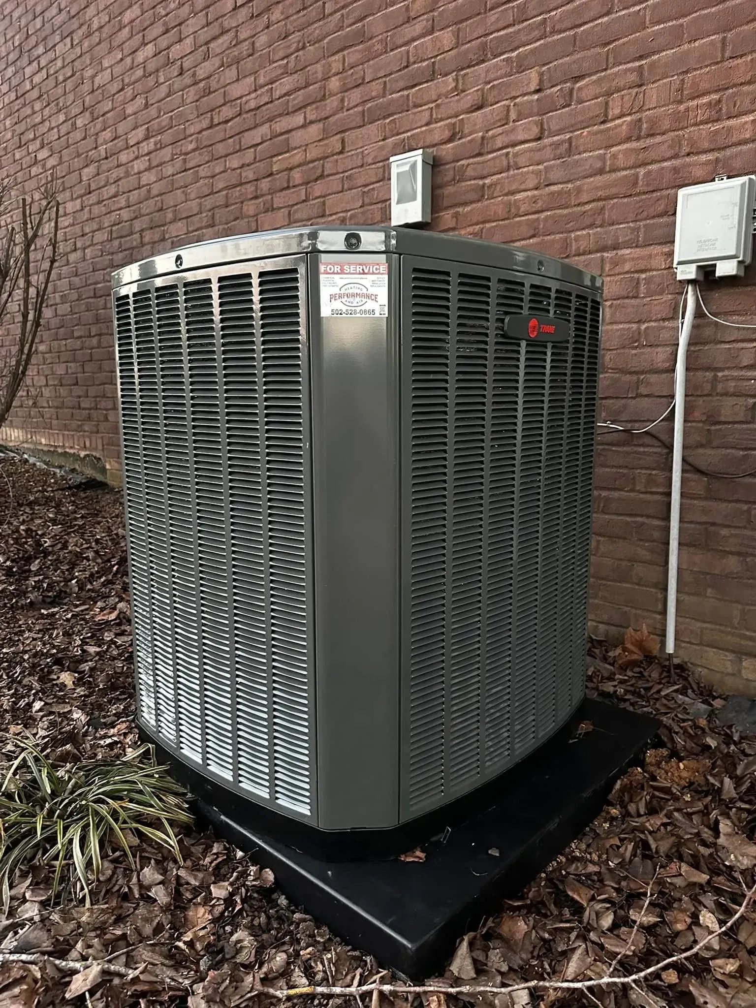 Outdoor HVAC unit on a black pad beside a brick wall, with leaves around it