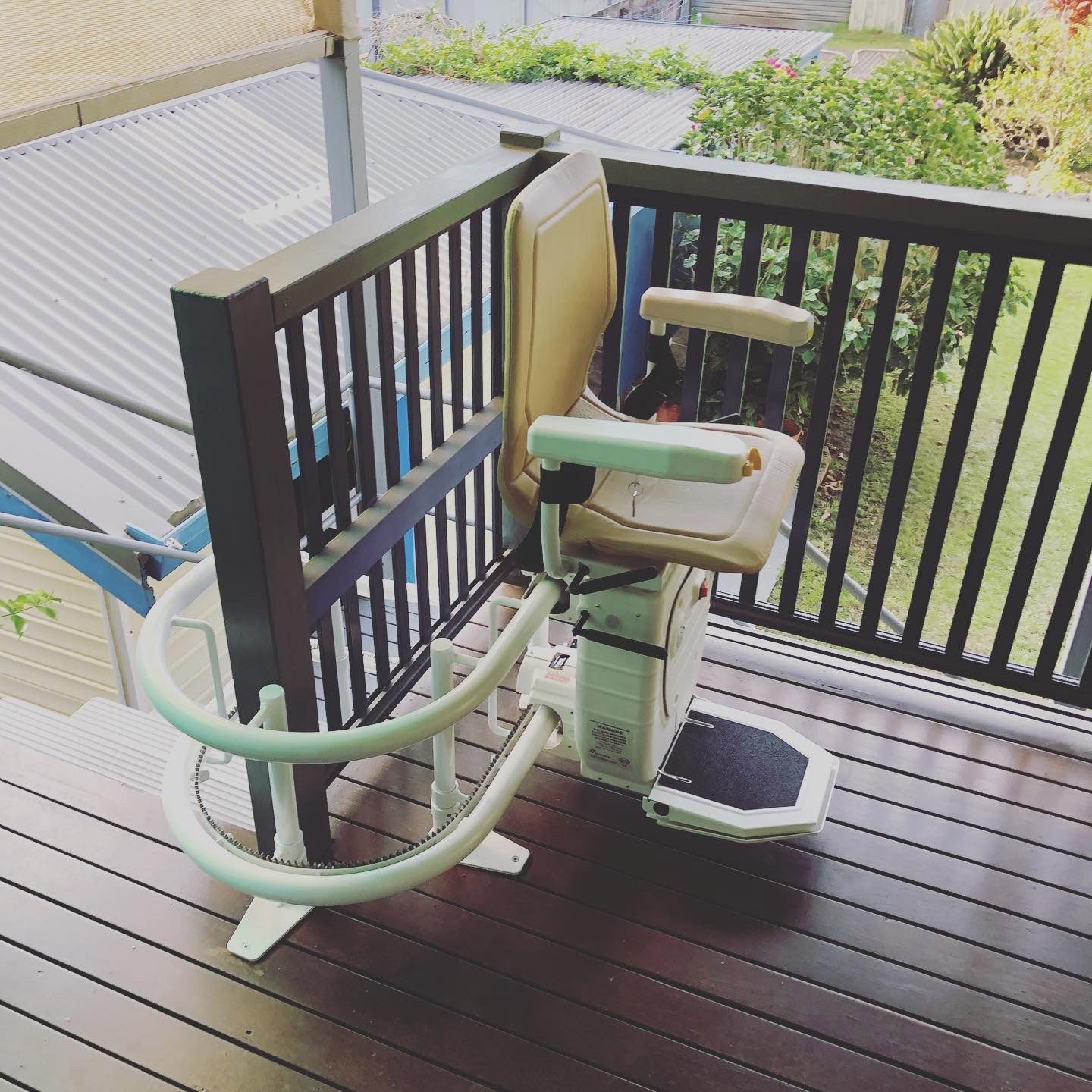 Stair lift on a wooden deck with a black railing, overlooking greenery. Lift is beige and white.