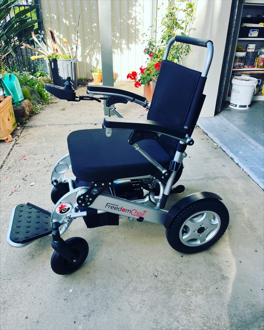 Silver and black electric wheelchair on a concrete surface outside a garage, with plants in the background.