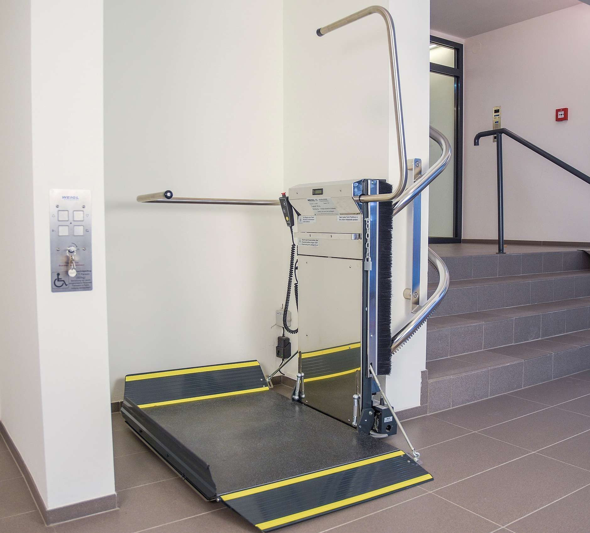 Stair lift platform attached to a wall, near stairs. Controls on the wall; ramp extended.