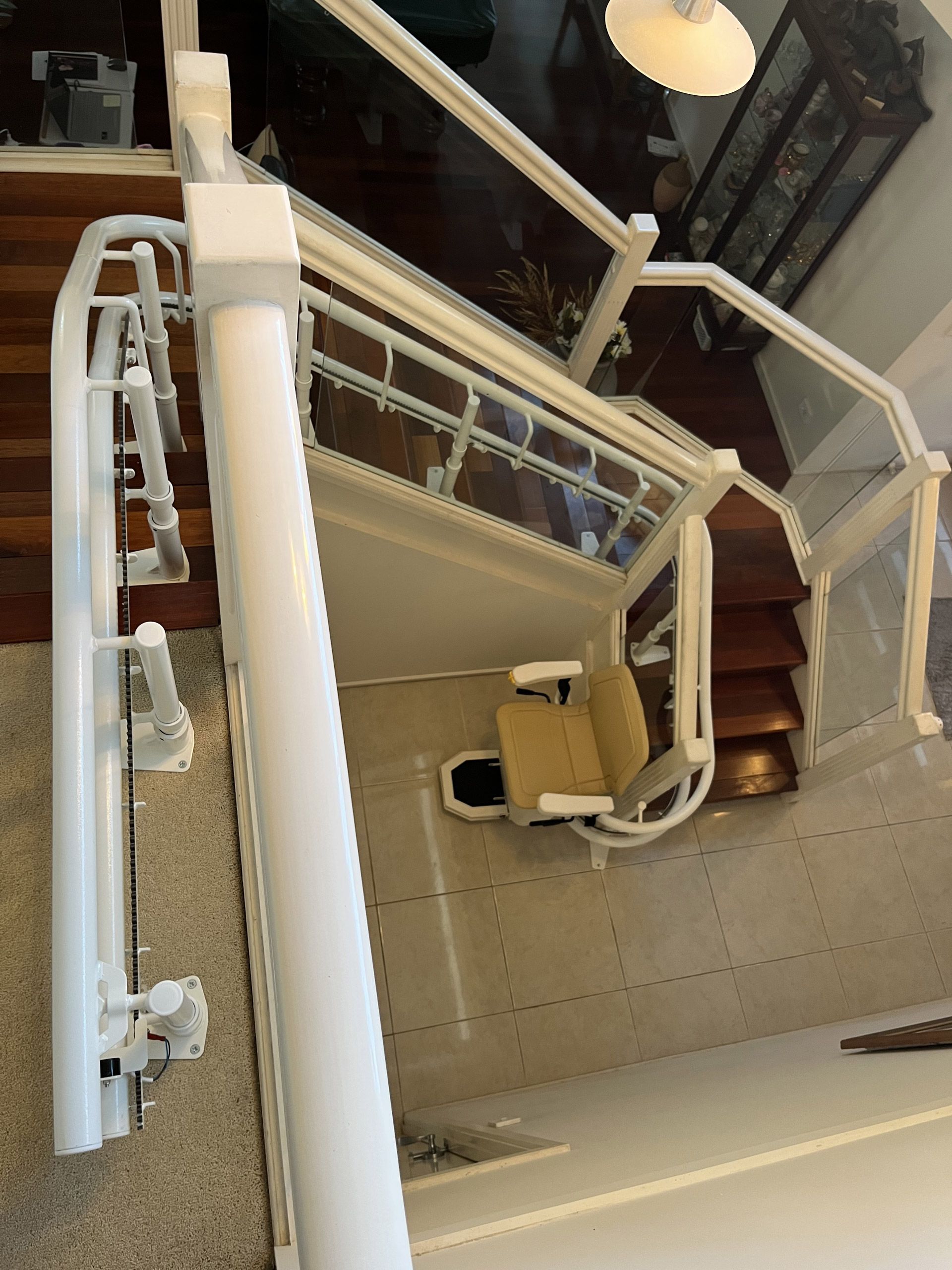 Stair lift on a wooden staircase with a beige seat. White railings with glass panels.