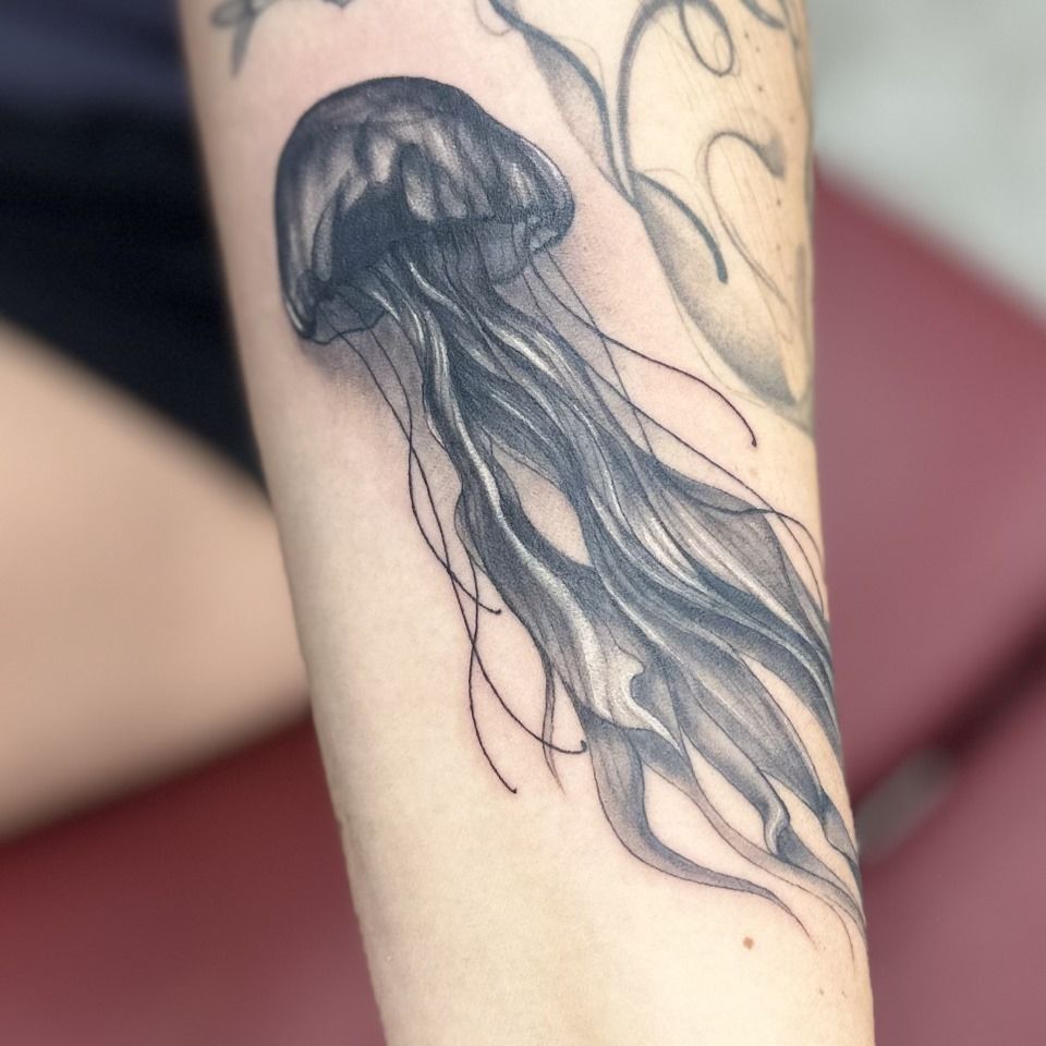 Black and gray jellyfish tattoo on a forearm; tentacles trail down.