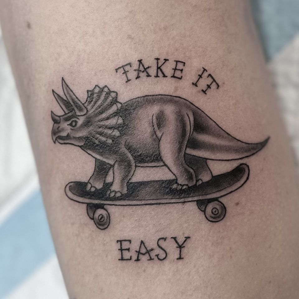 Tattoo of a triceratops riding a skateboard with text 