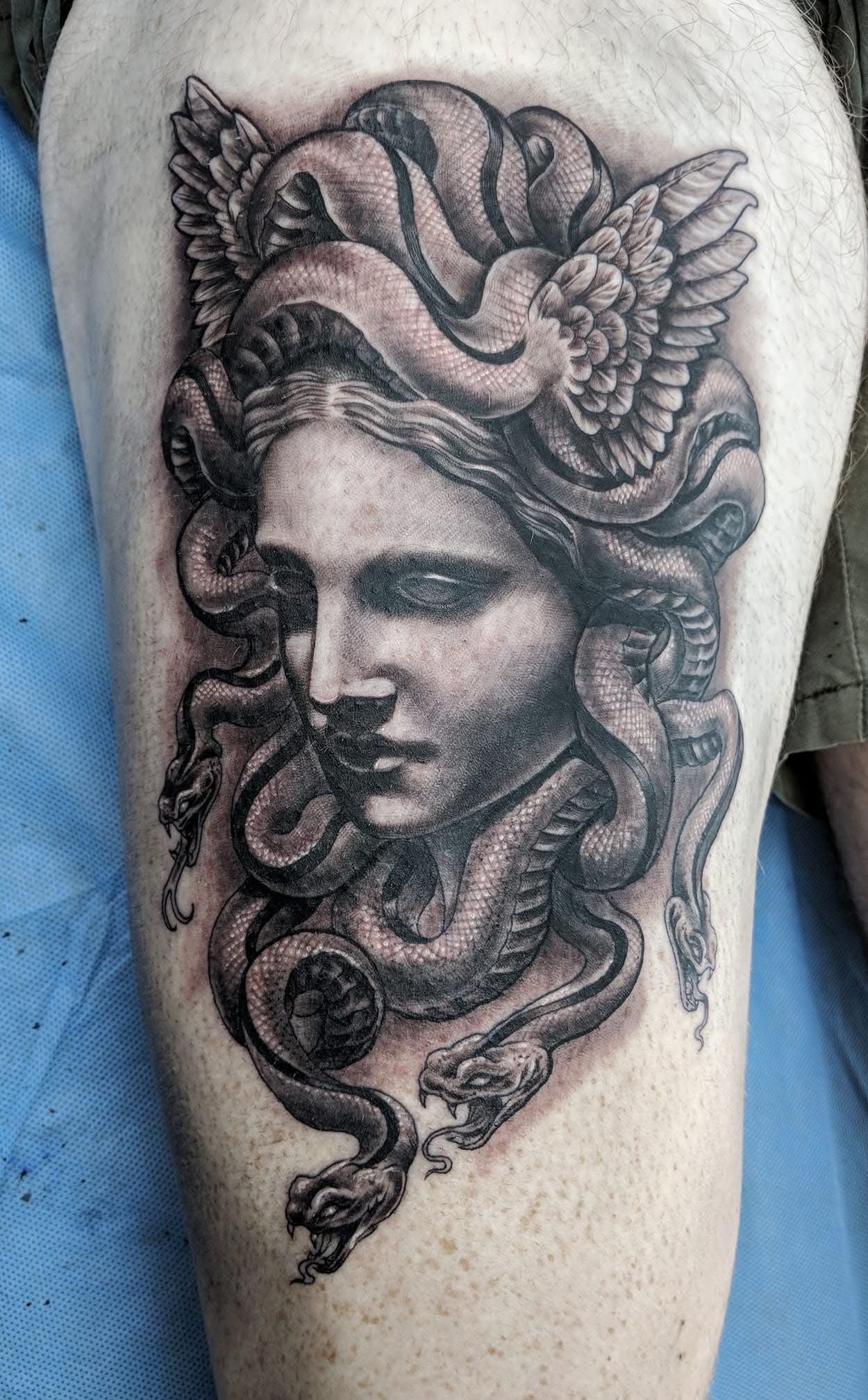 Tattoo of Medusa's head with flowing snakes and winged head covering, on a thigh. Black and gray shading. — Whitsunday Tattooing Airlie Beach in Whitsundays, QLD