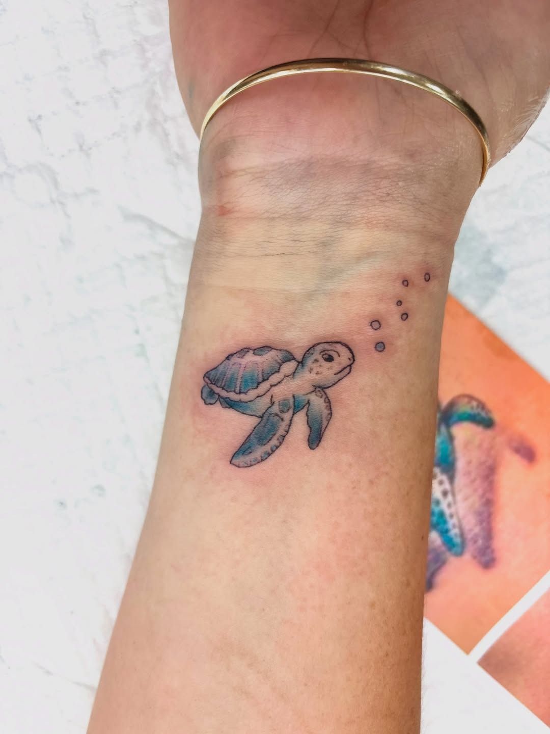 Hand with a small fish tattoo on the wrist, beside a blue and orange tattoo design in the background — Whitsunday Tattooing Airlie Beach in Whitsundays, QLD