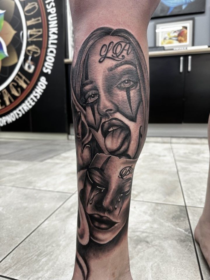 Black and gray tattoo of two faces on a leg. One face sticks out tongue, and the other cries. 