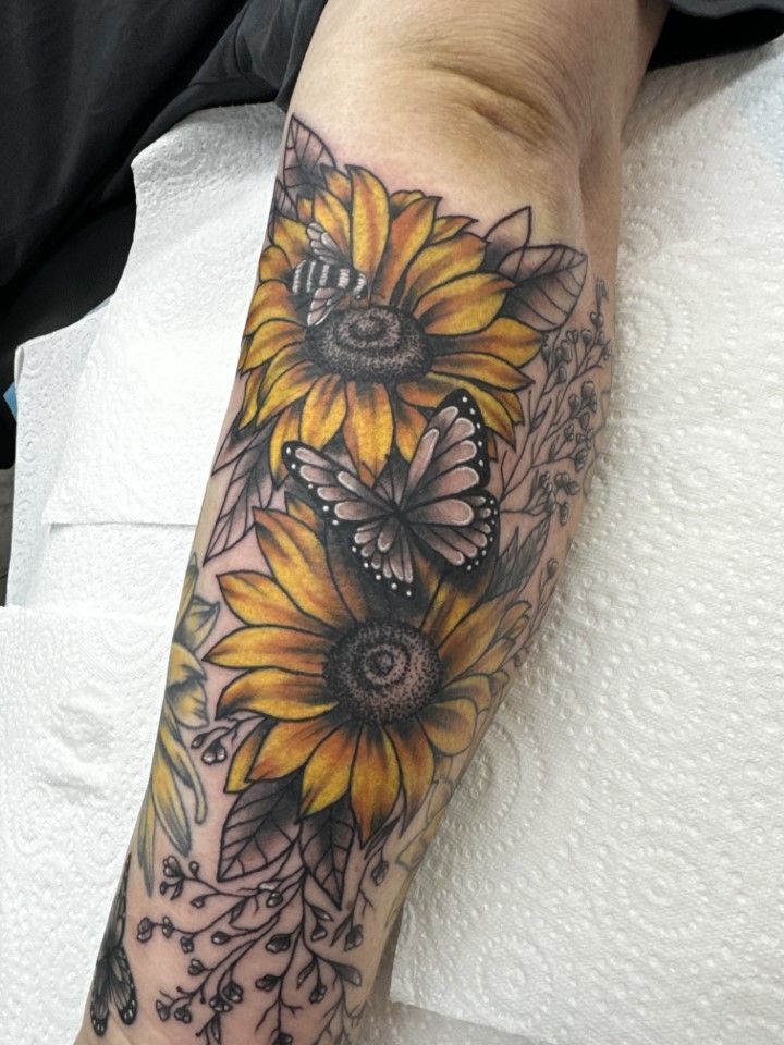 Tattoo of two sunflowers and a butterfly on an arm. Yellow, black, and gray ink.