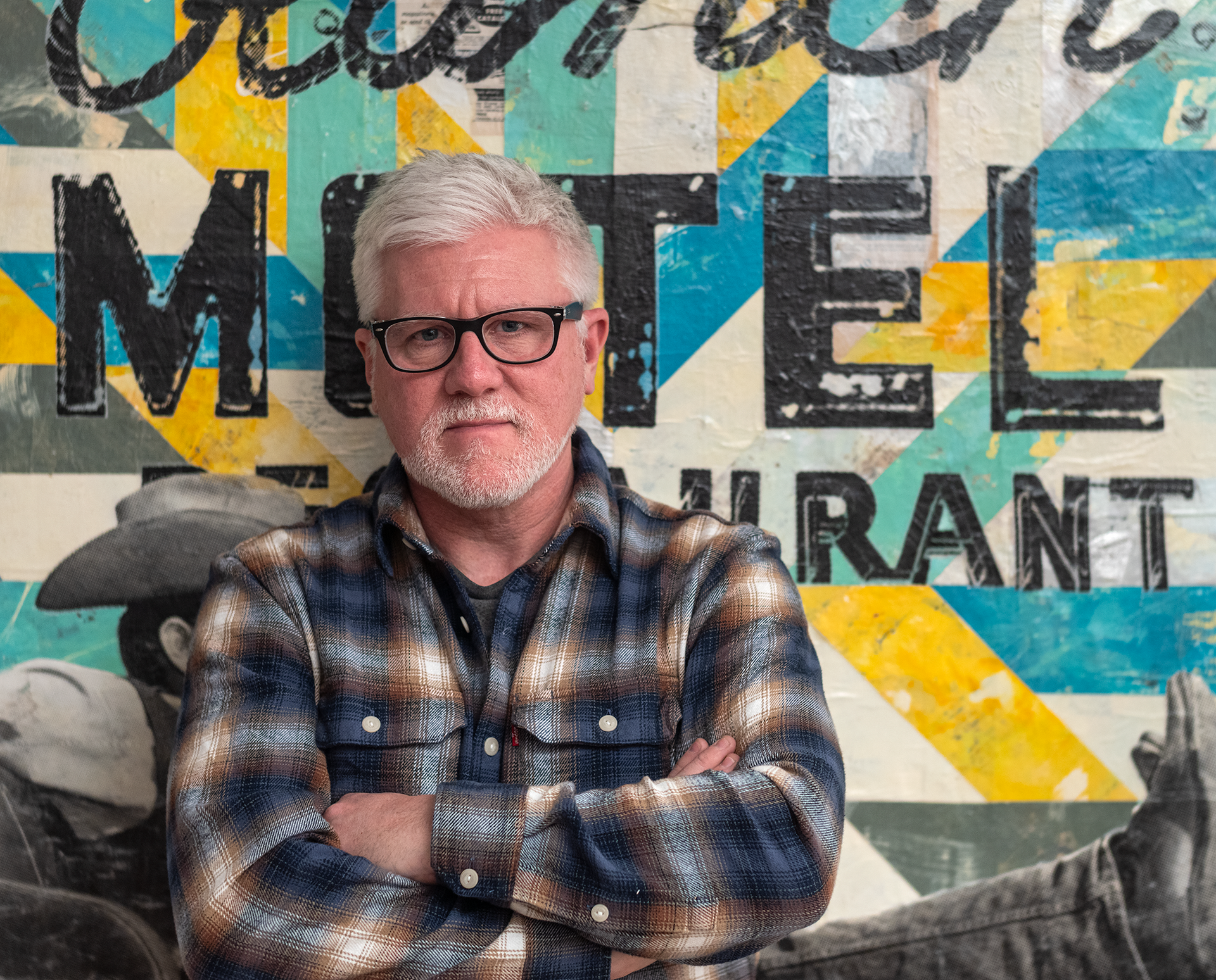 Artist Robert Mars with white hair and glasses in a plaid shirt stands with arms crossed before a colorful art background.