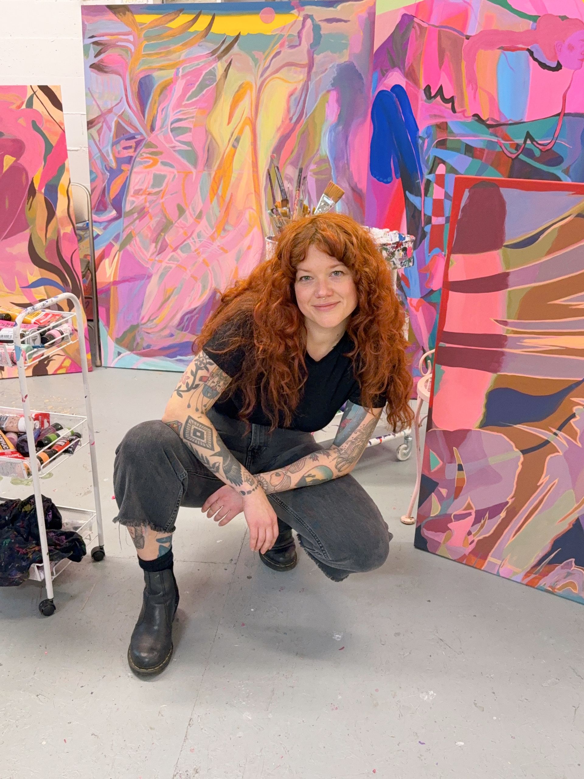 Artist Emily Wise with long red hair and arm tattoos crouches in an art studio with several large colorful abstract canvases.