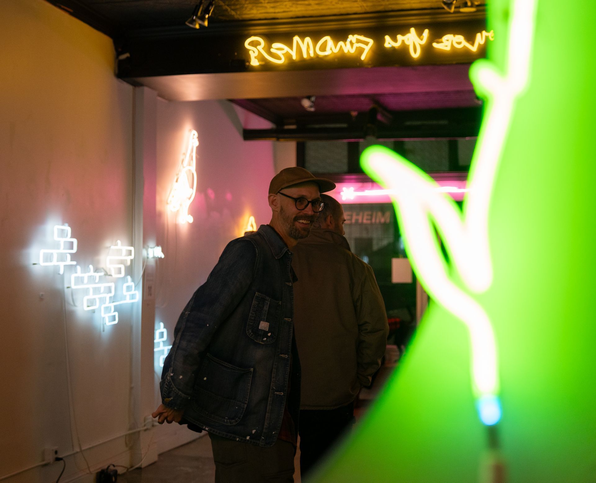 Yale Wolf in a denim jacket and glasses smiles inside a gallery space filled with glowing neon art and wall lights.